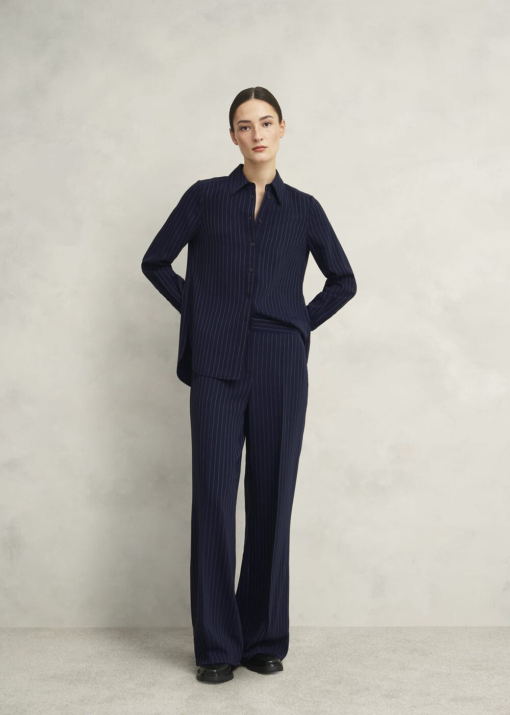 A woman wearing navy wide-leg trousers with a pinstripe pattern, a matching navy pinstripe shirt, and black shoes, standing against a plain background.