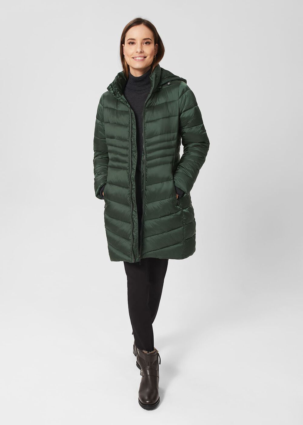 A woman wearing a green Petite Nessie Puffer jacket with a detachable hood, fleece-lined pockets, and ribbed cuffs, standing against a plain background.