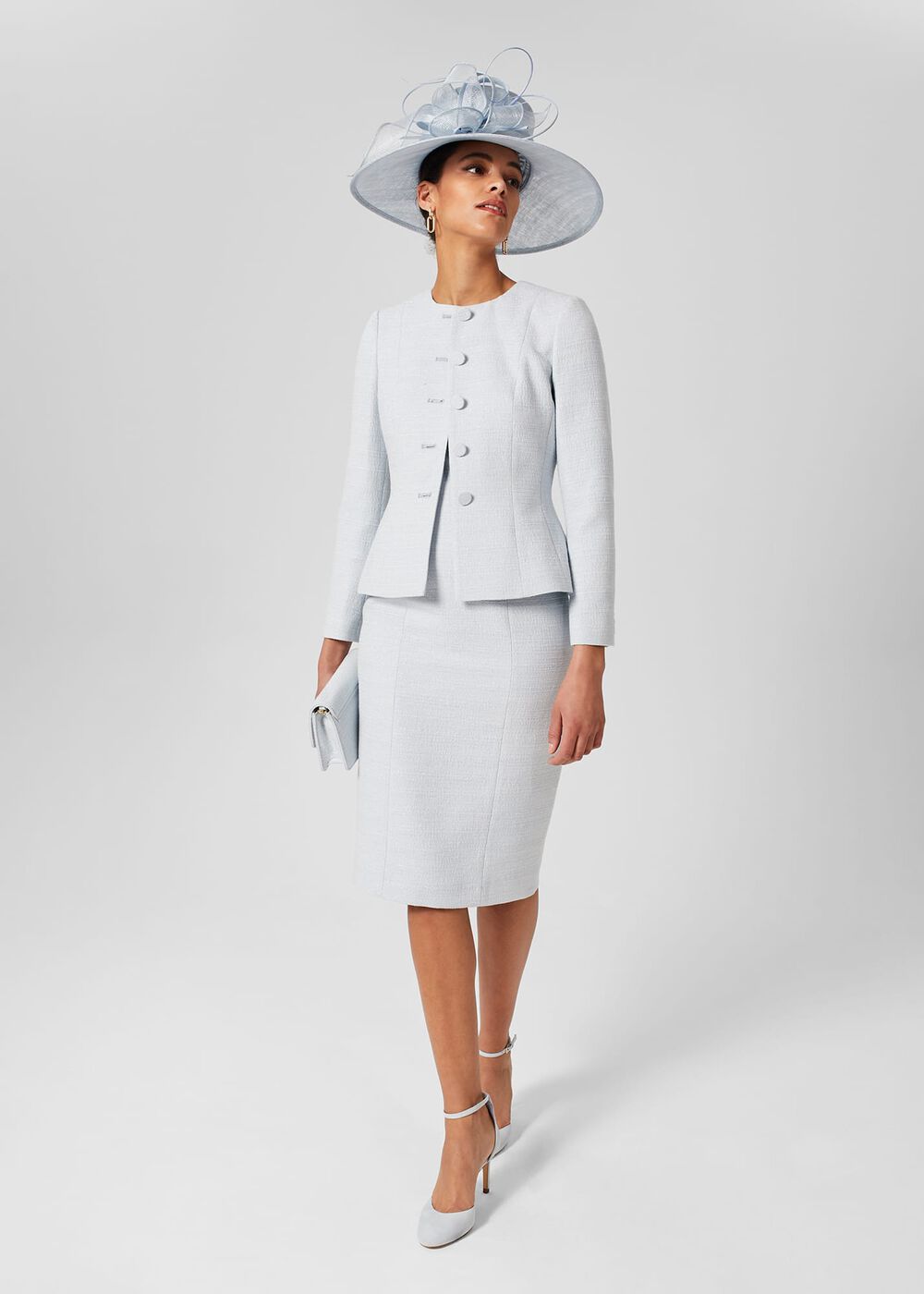 A woman wearing a white textured tailored jacket with a peplum shape, paired with a matching skirt, accessorized with earrings and a large hat.
