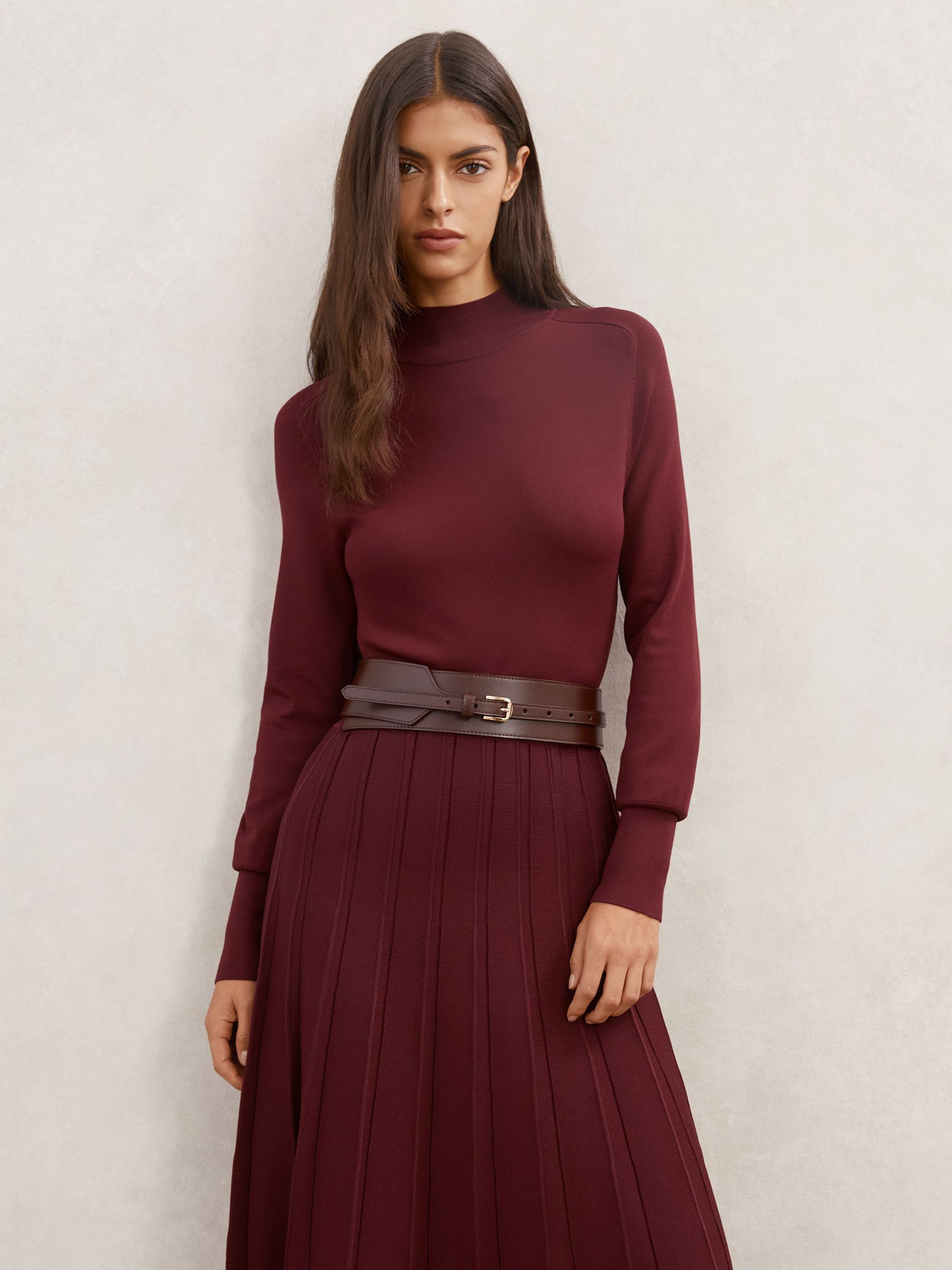 A woman wearing a Raspberry Red midi dress with a high neckline, saddle-style sleeves, a wide leather belt, and a pleated skirt.