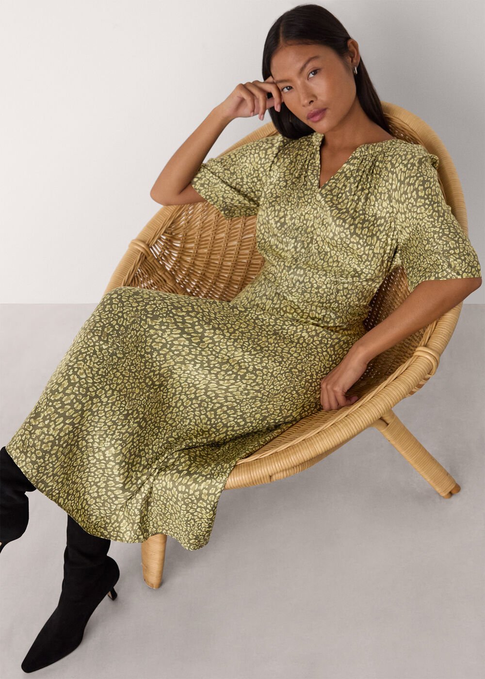 A woman wearing a green leopard print midi dress with a V-neckline, tie belt at the waist, and side split, sitting in a wicker chair.