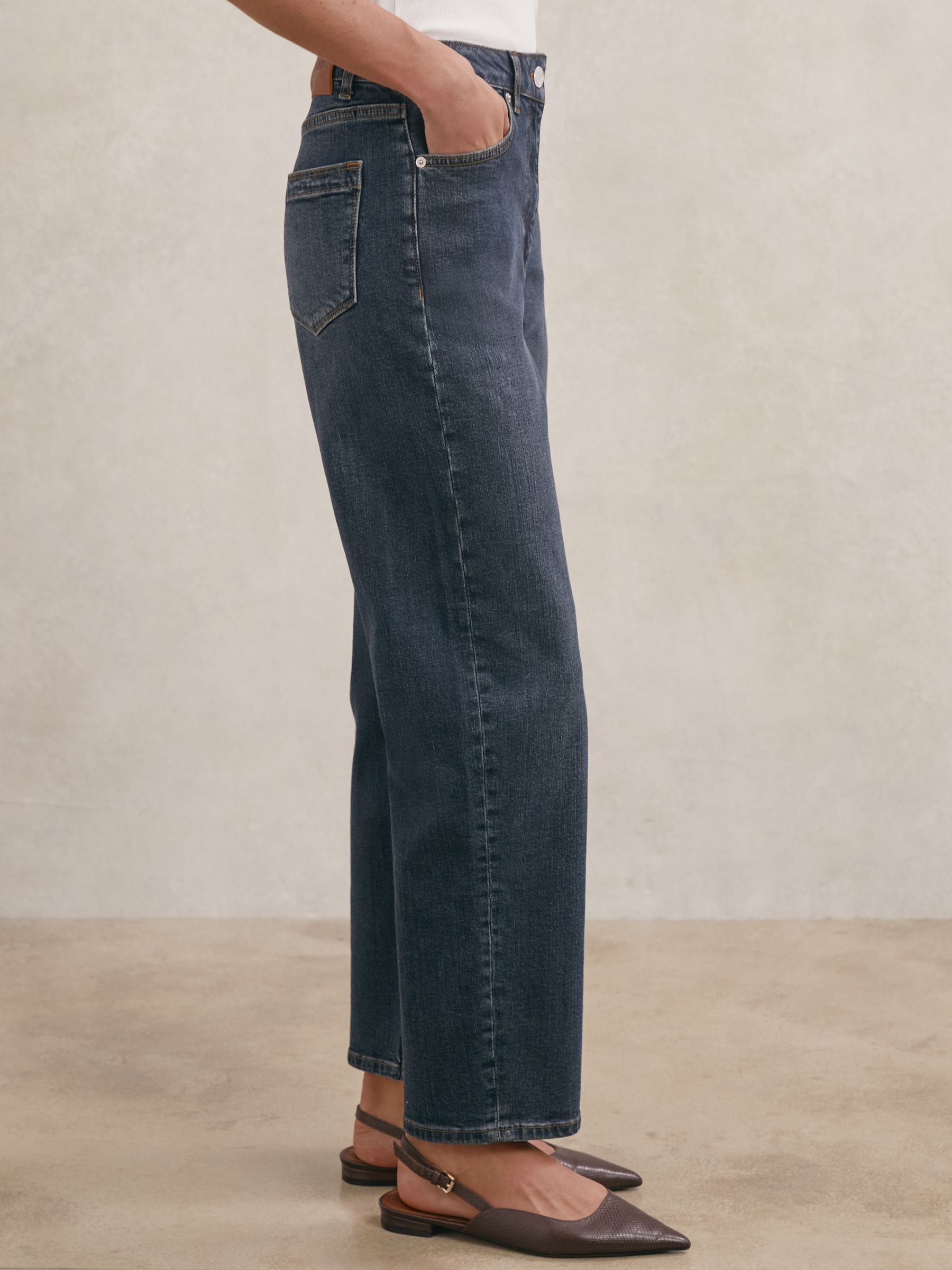 A pair of petite mid-rise straight-leg jeans in antique mid blue denim, with five pockets, a front button and zip closure, worn by a person with their hand in the pocket.