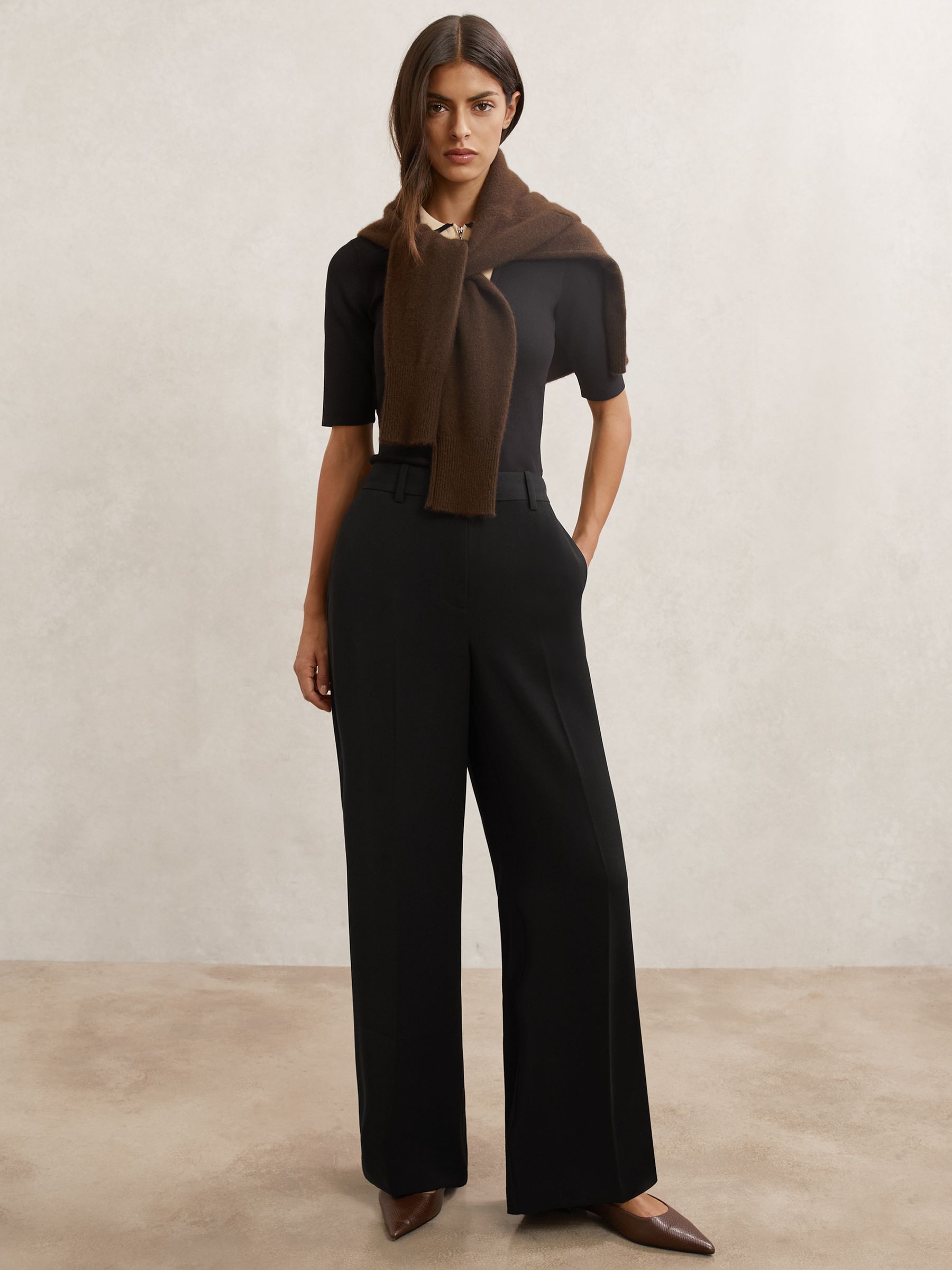 A woman wearing black wide-leg tailored trousers with a fluid fabric, pressed creases, and functional pockets, paired with a black top and brown shoes.