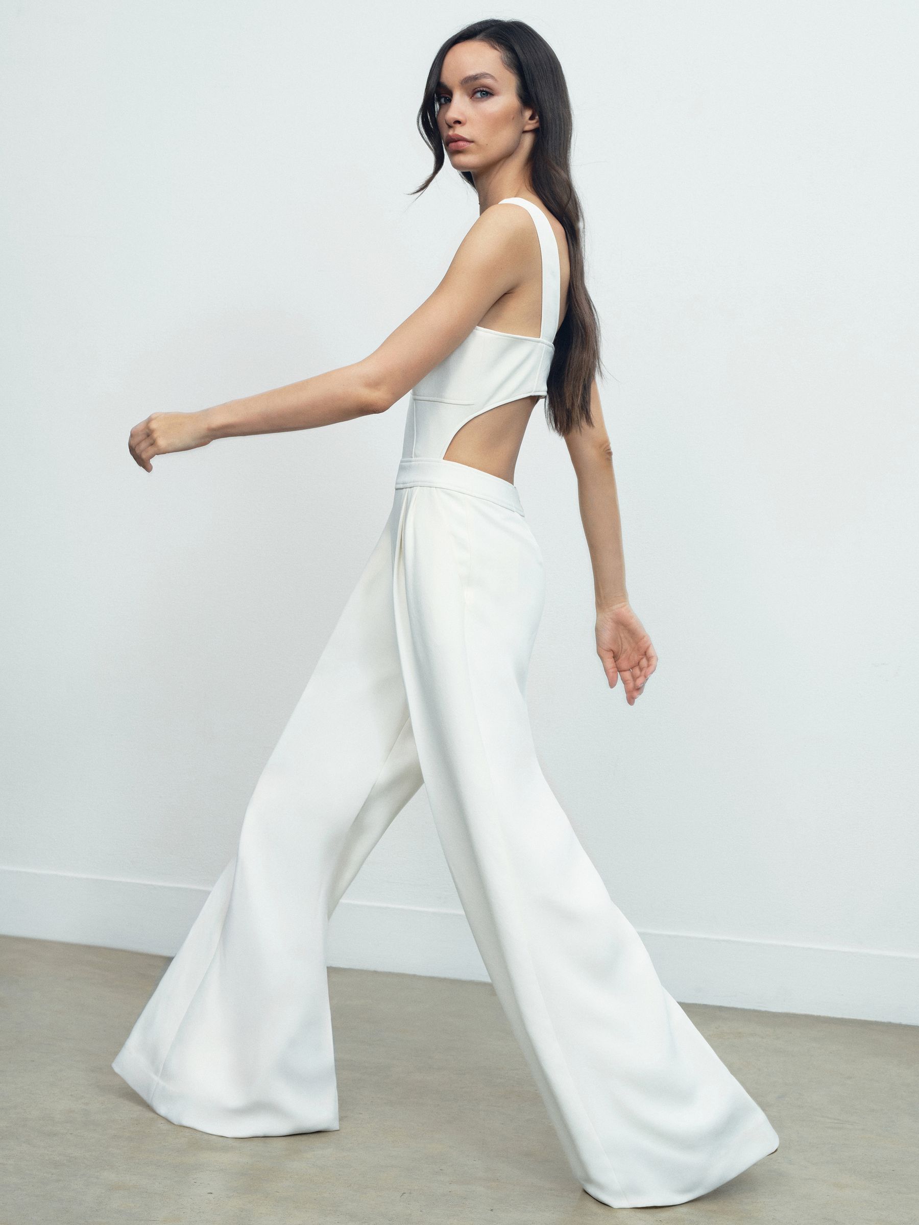 A woman wearing an ivory wide-leg jumpsuit with cut-outs at the sides, square neckline, and pleated front, standing against a plain background.