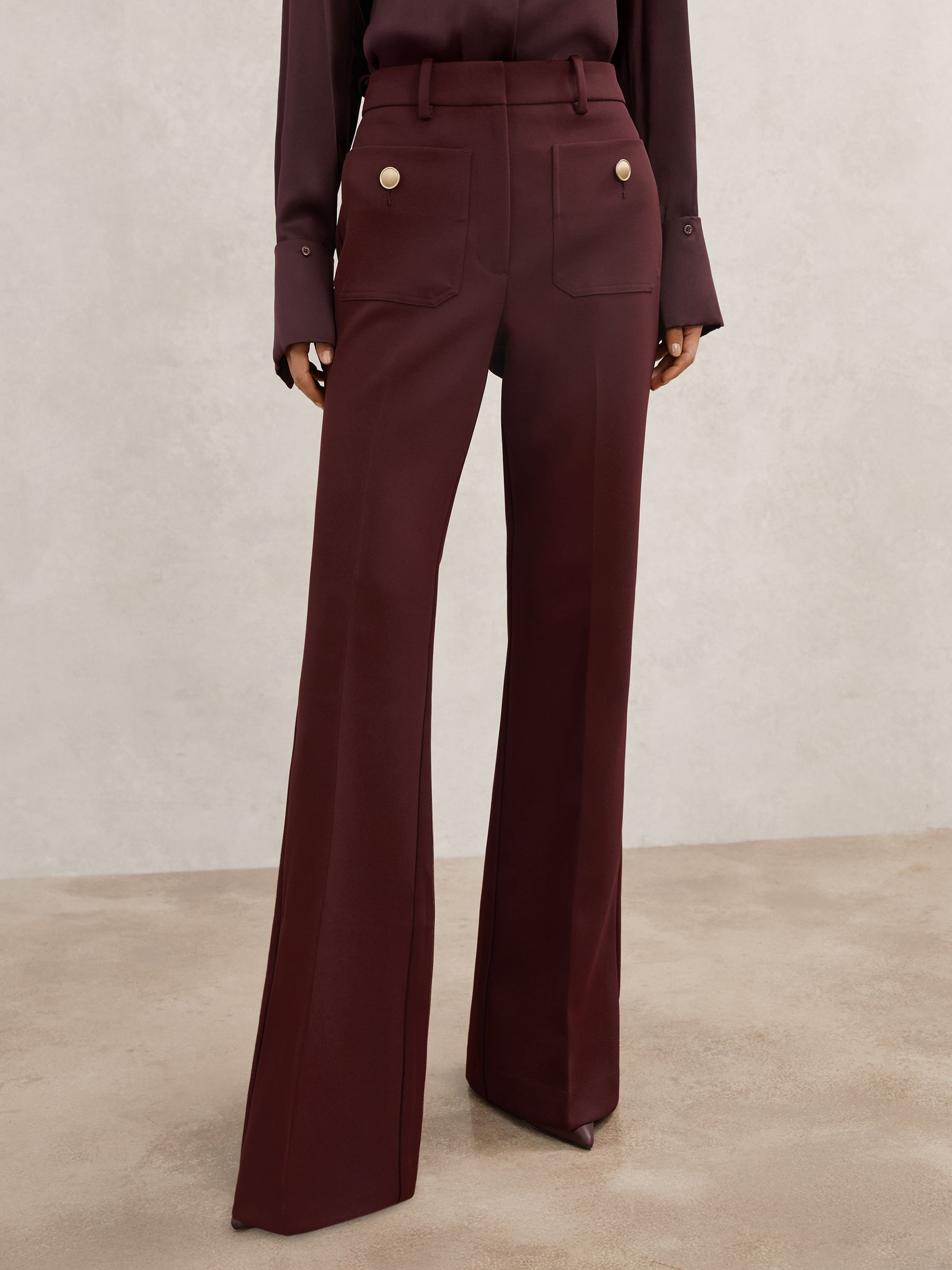 A pair of dark red petite flared trousers with twin patch pockets and gold-tone buttons, shown on a model.