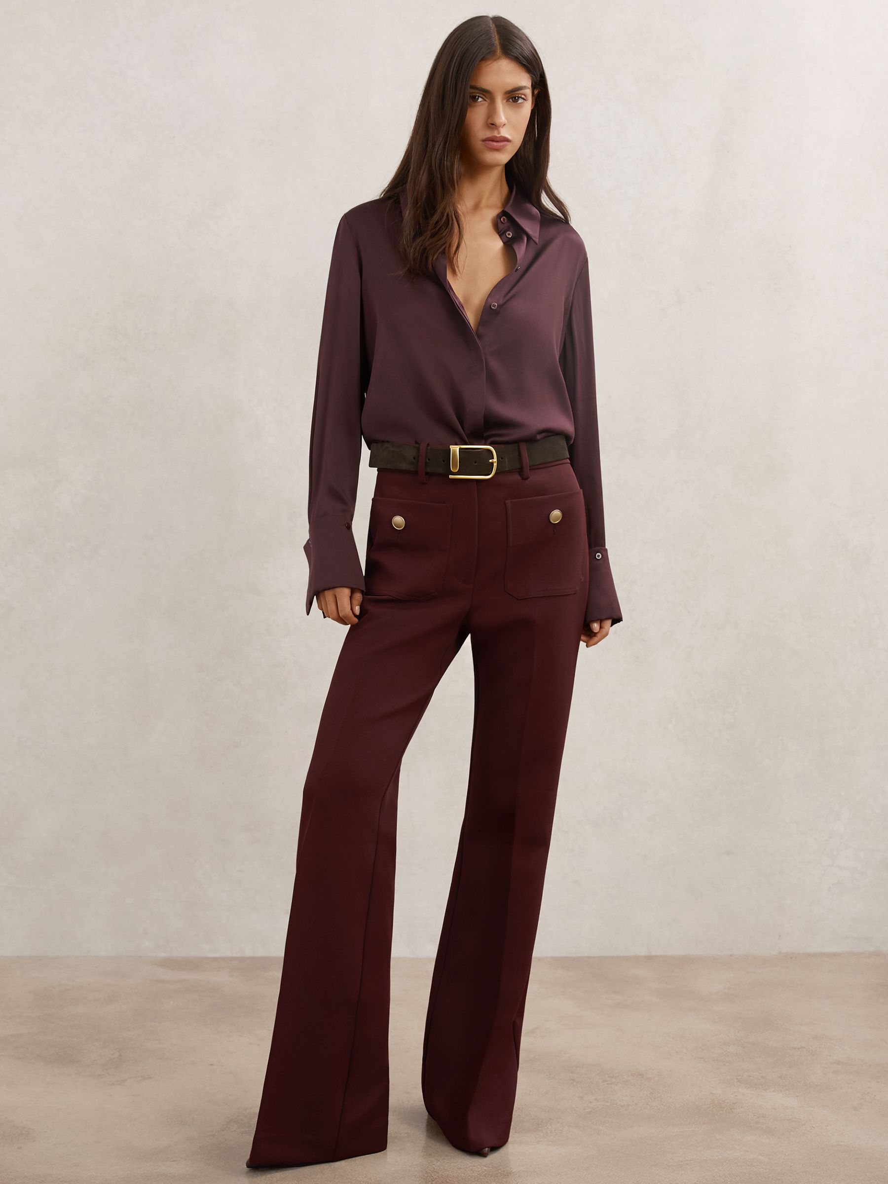 A woman wearing dark red flared trousers with patch pockets, gold buttons, and a zip closure, paired with a matching dark red blouse.