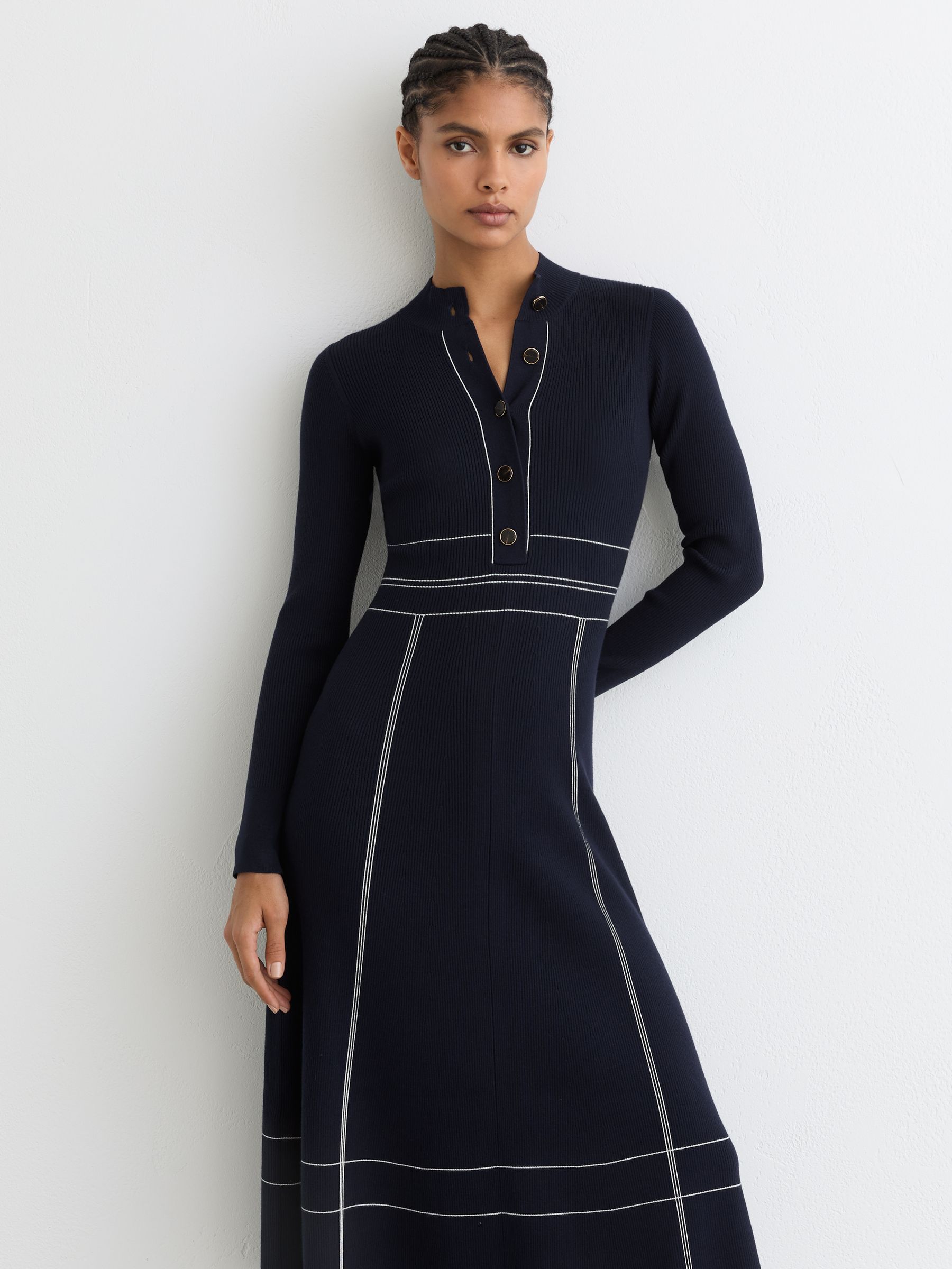 A woman wearing a navy knitted midi dress with contrast stitching, high neckline, and a half-button placket, standing against a white wall.