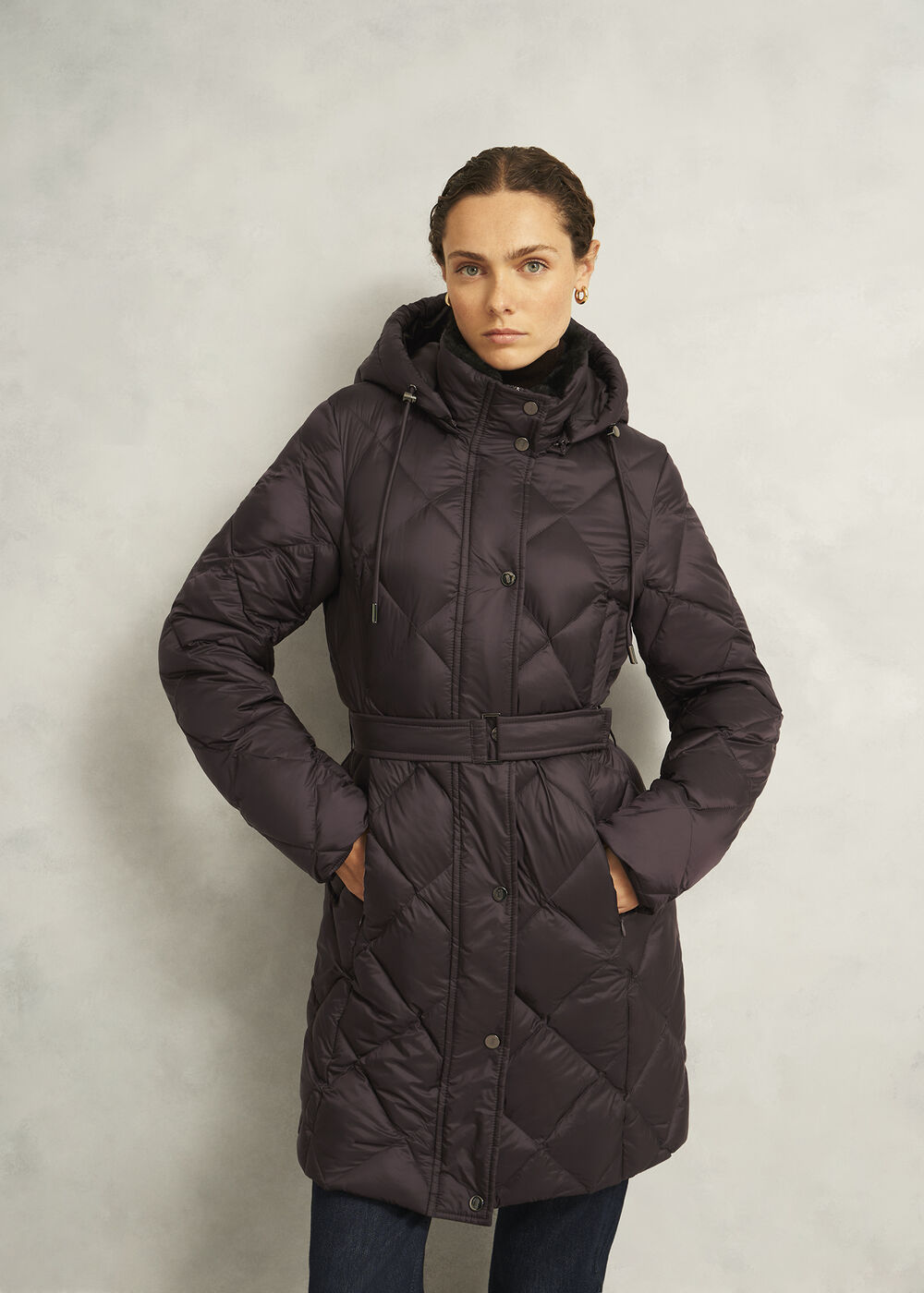A woman wearing a dark purple quilted puffer coat with a hood, faux fur collar, and buckle belt, standing against a plain background.