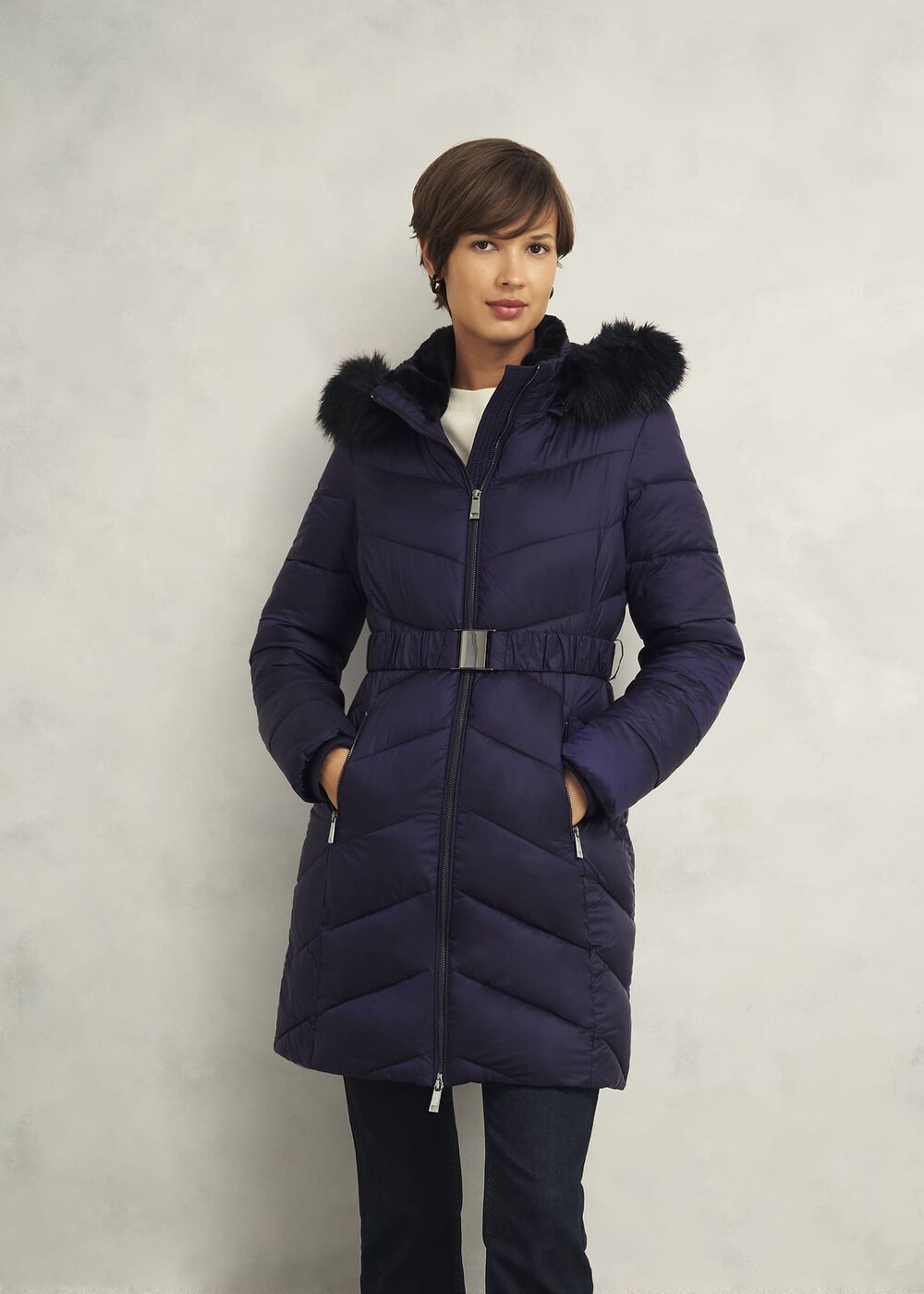 A woman wearing a navy blue puffer coat with a hood and black faux fur trim, front zipper, and belted waist, standing against a plain background.