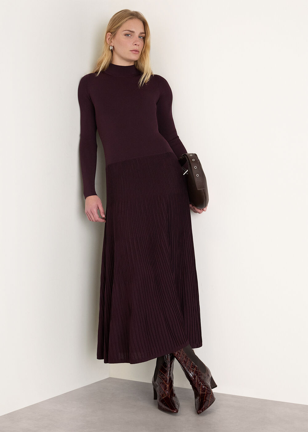 A woman wearing a plum-colored knit dress with a funnel neck and pleated A-line skirt, paired with patterned high-heeled boots and holding a dark handbag.