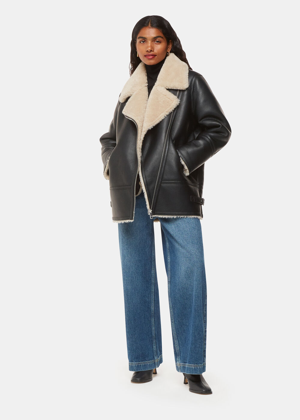 A woman wearing a black petite faux fur aviator jacket with a faux shearling collar, zip closure, and zipped pockets, paired with blue jeans and black boots.