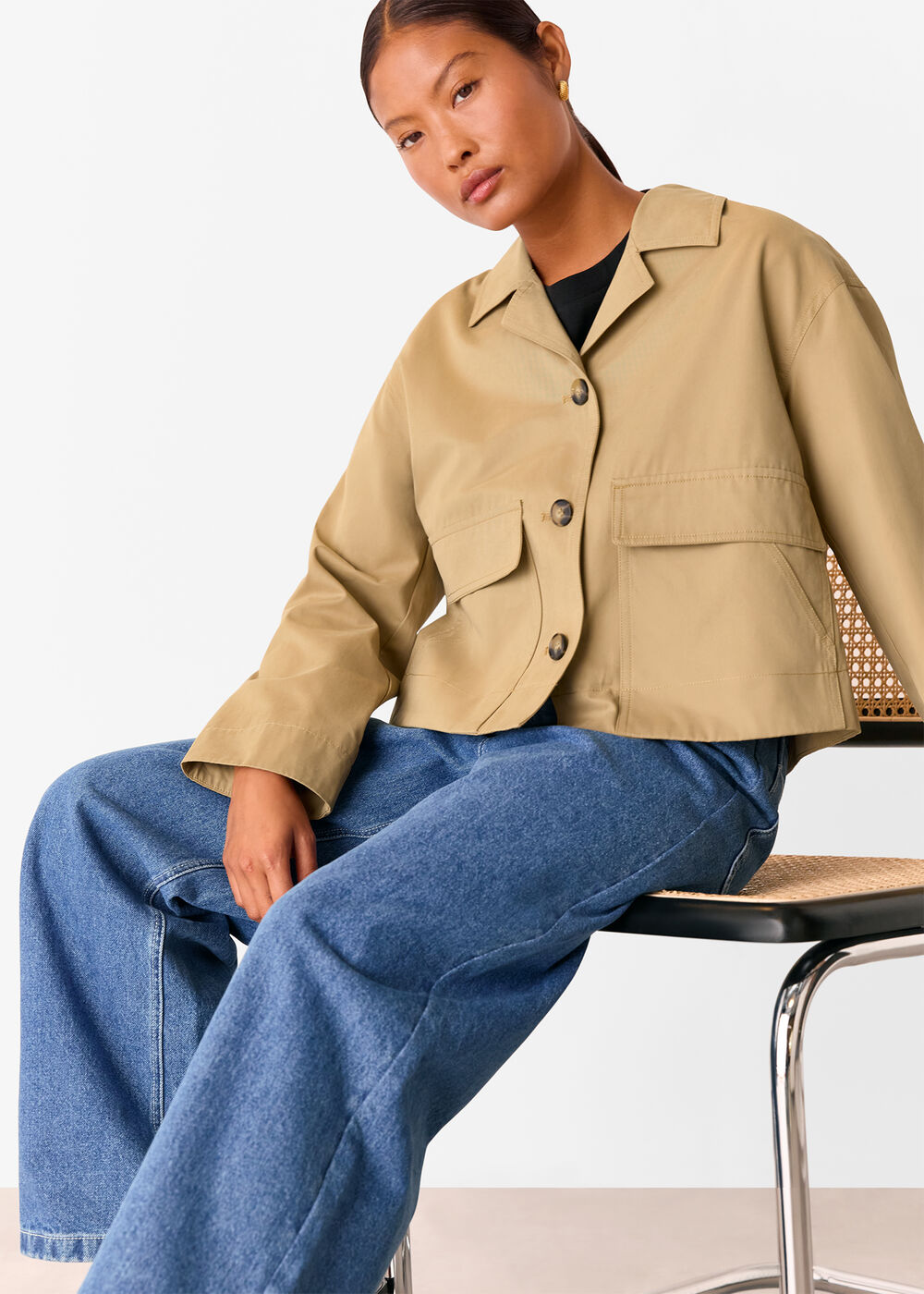 A woman wearing a beige, short cotton trench jacket with a collar, large front patch pockets, and a back vent, paired with blue jeans.