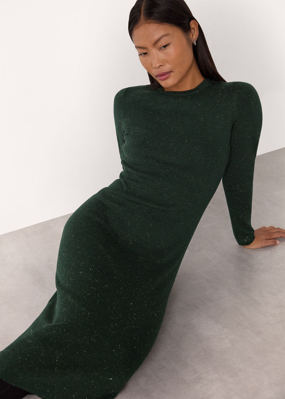 A woman wearing a dark green, long-sleeved, bodycon knit dress with a round neckline and subtle metallic sparkle, sitting on a light-colored surface.
