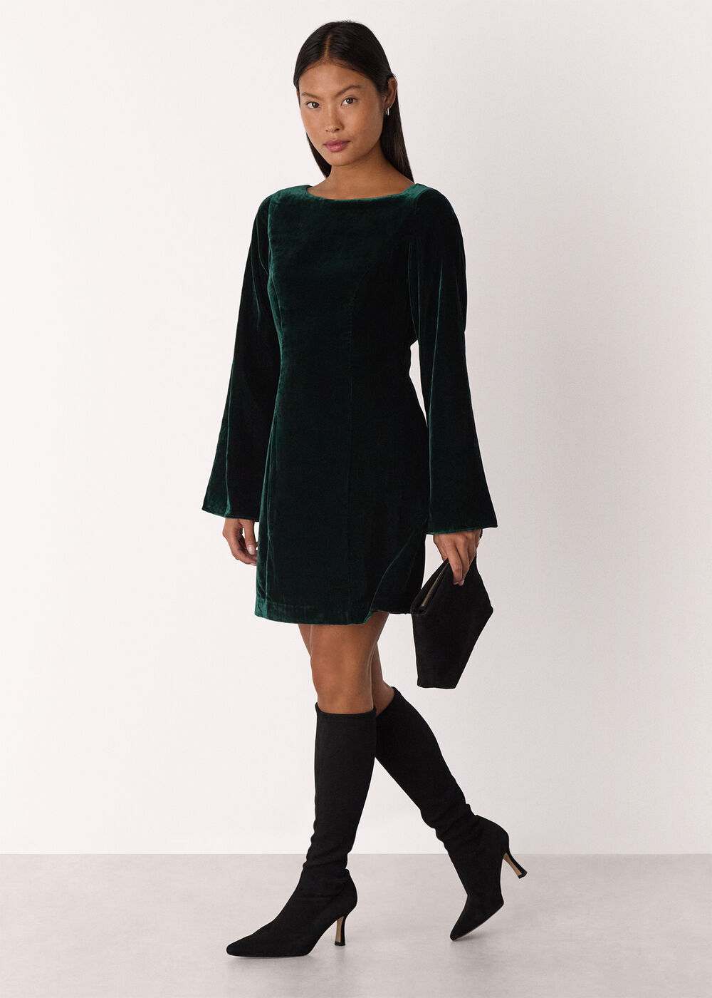 A woman wearing a forest green velvet dress with flare sleeves, square neckline, and a back keyhole detail, paired with black knee-high boots and holding a black clutch.