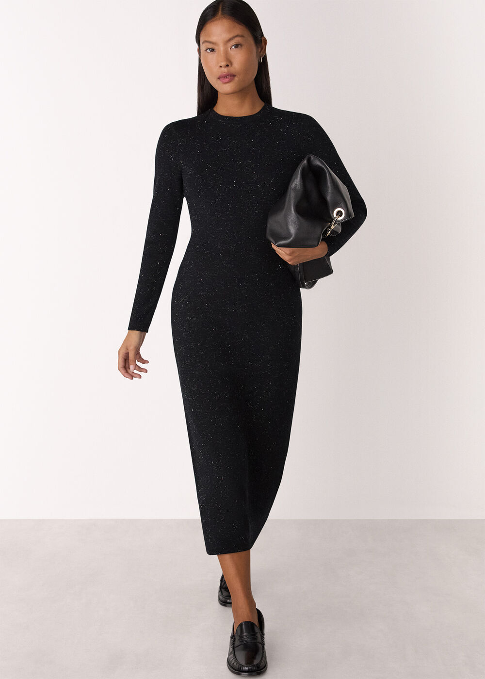A woman wearing a black, long-sleeved, metallic fiber knit dress with a crew neck and midi length, holding a black bag, standing against a plain background.