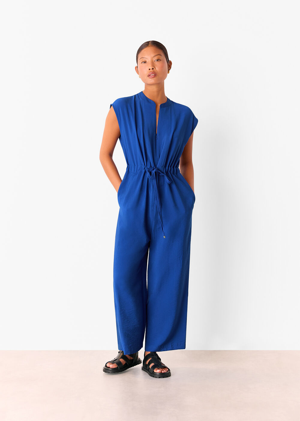 A woman wearing a cobalt blue sleeveless jumpsuit with a V-neckline and a drawstring waist, paired with black sandals.