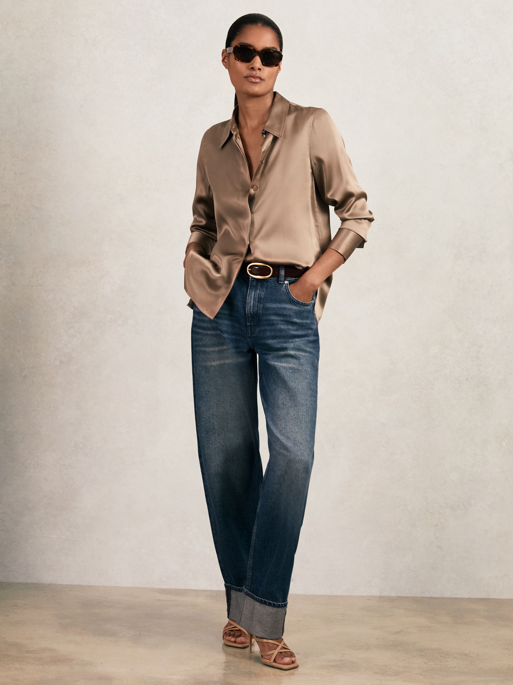 A woman wearing a taupe brown silk satin shirt with a pointed collar and concealed placket, paired with blue jeans and sunglasses.