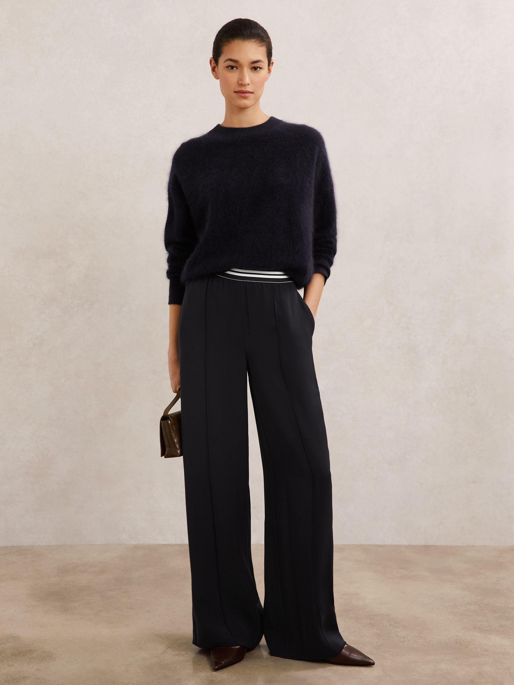 A woman wearing navy wide-leg trousers with an elasticated waistband, front pintucks, and side pockets, paired with a dark sweater and brown shoes.