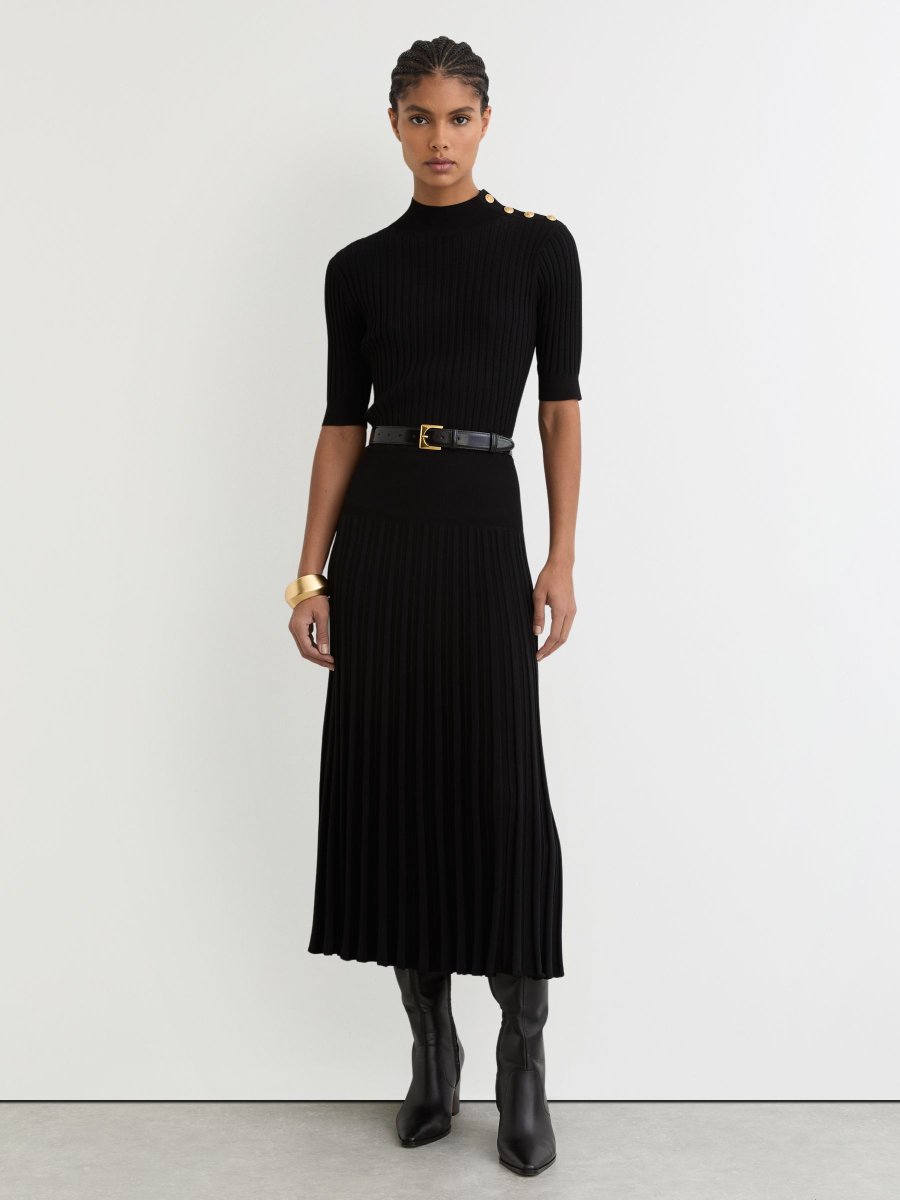 A woman wearing a black knitted midi dress with a high neckline, short sleeves, crest-embossed press-studs on the shoulder, and a pleated skirt.