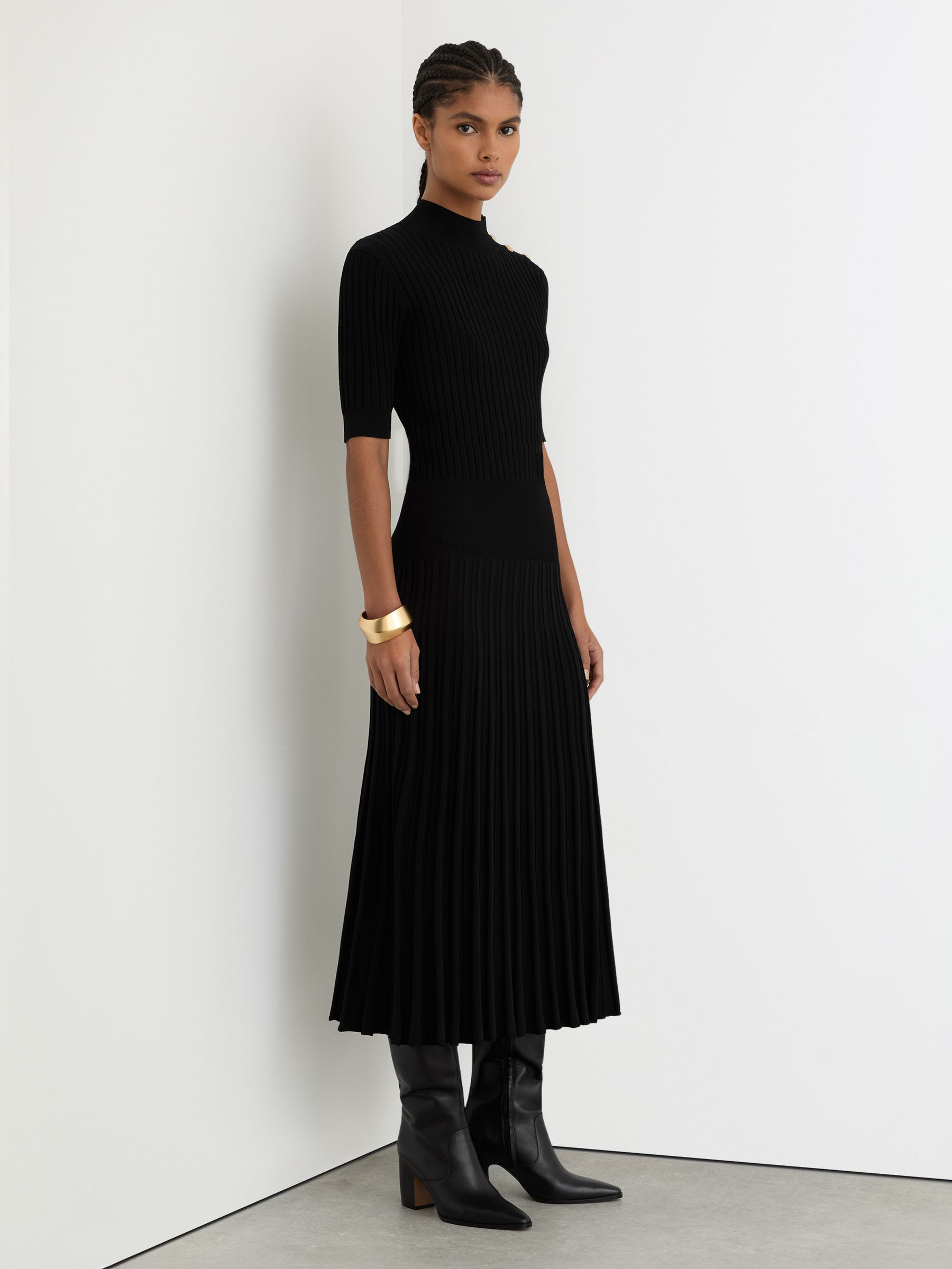 A woman wearing a black knitted midi dress with a high neckline, short sleeves, crest-embossed press-studs on the shoulder, a pleated skirt, and textured perforated rib-knit details.
