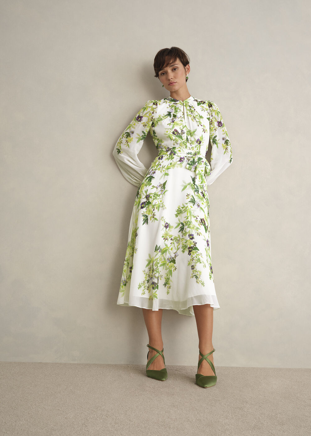 A woman wearing a white silk floral dress with green and purple floral patterns, long sleeves, and a tie waist, standing against a plain background.