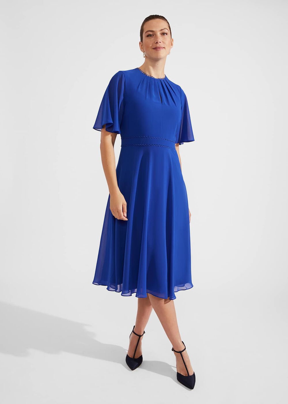 A woman wearing a royal blue fit and flare midi dress with short frill sleeves, gathered crew neckline, and a fitted waist, paired with black high heels.