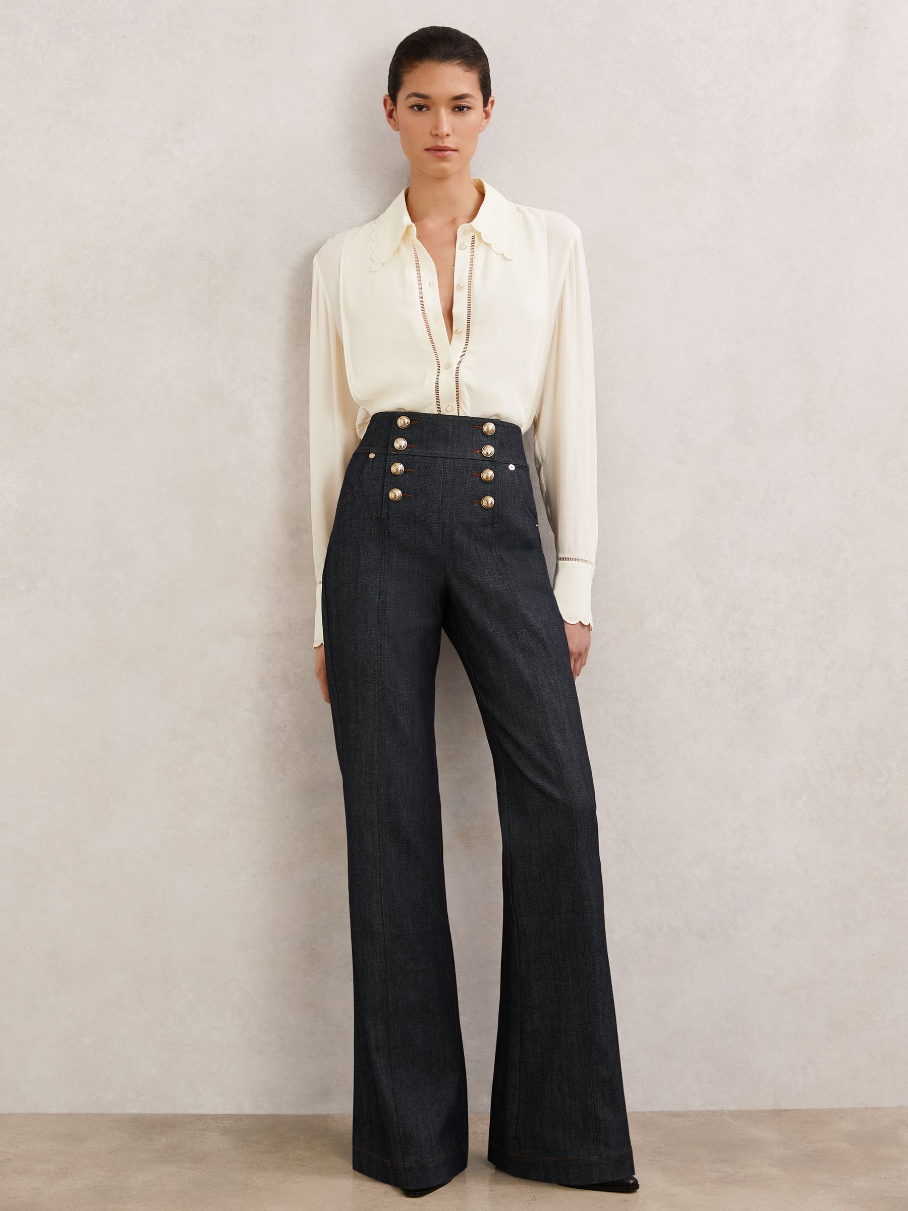 A woman wearing dark blue flared jeans with crest-embossed buttons, side and back pockets, paired with a cream blouse.
