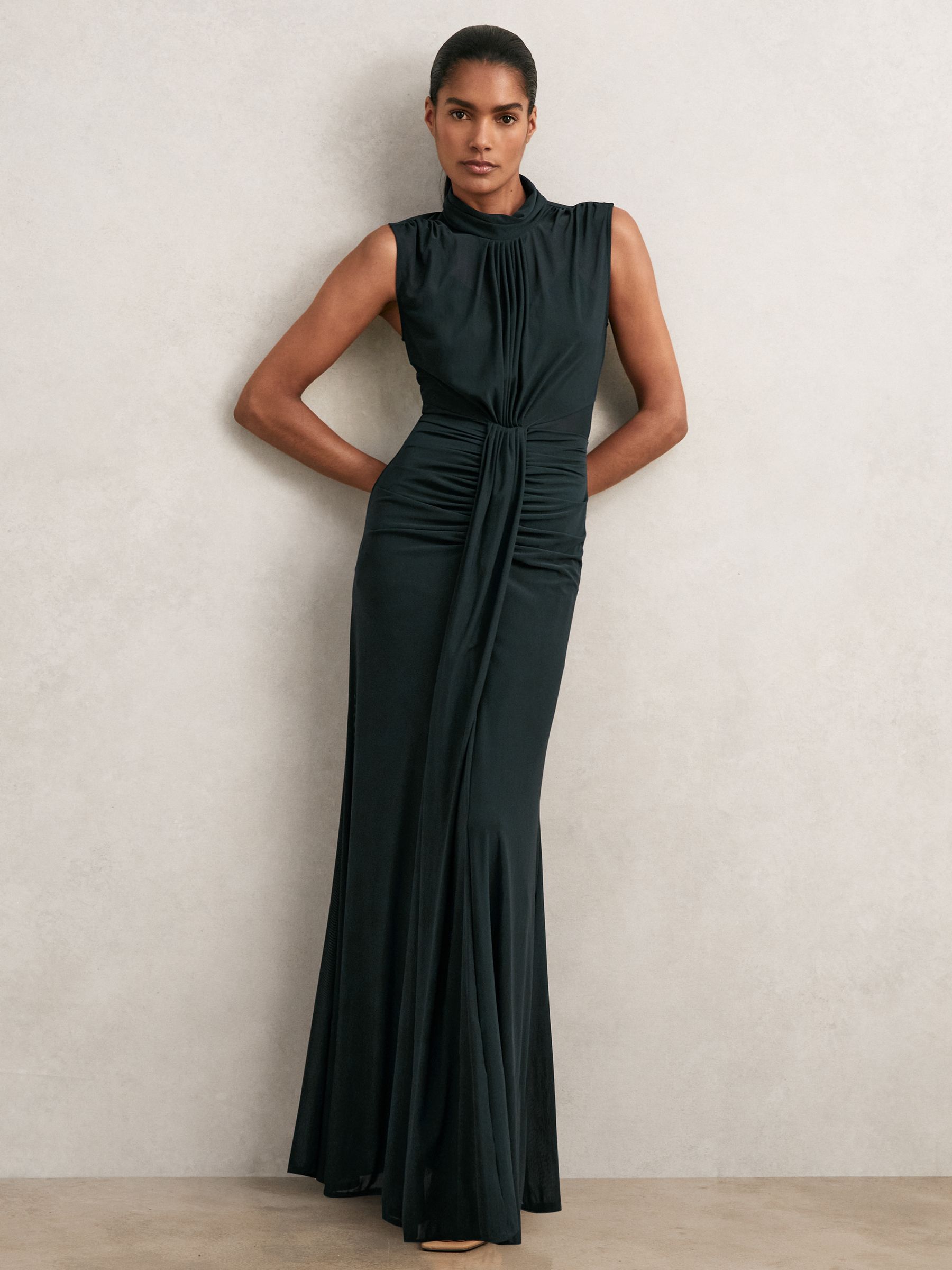 A woman wearing a sleeveless teal maxi dress with a high neckline, sheer fabric, ruching details, and a flared hem.