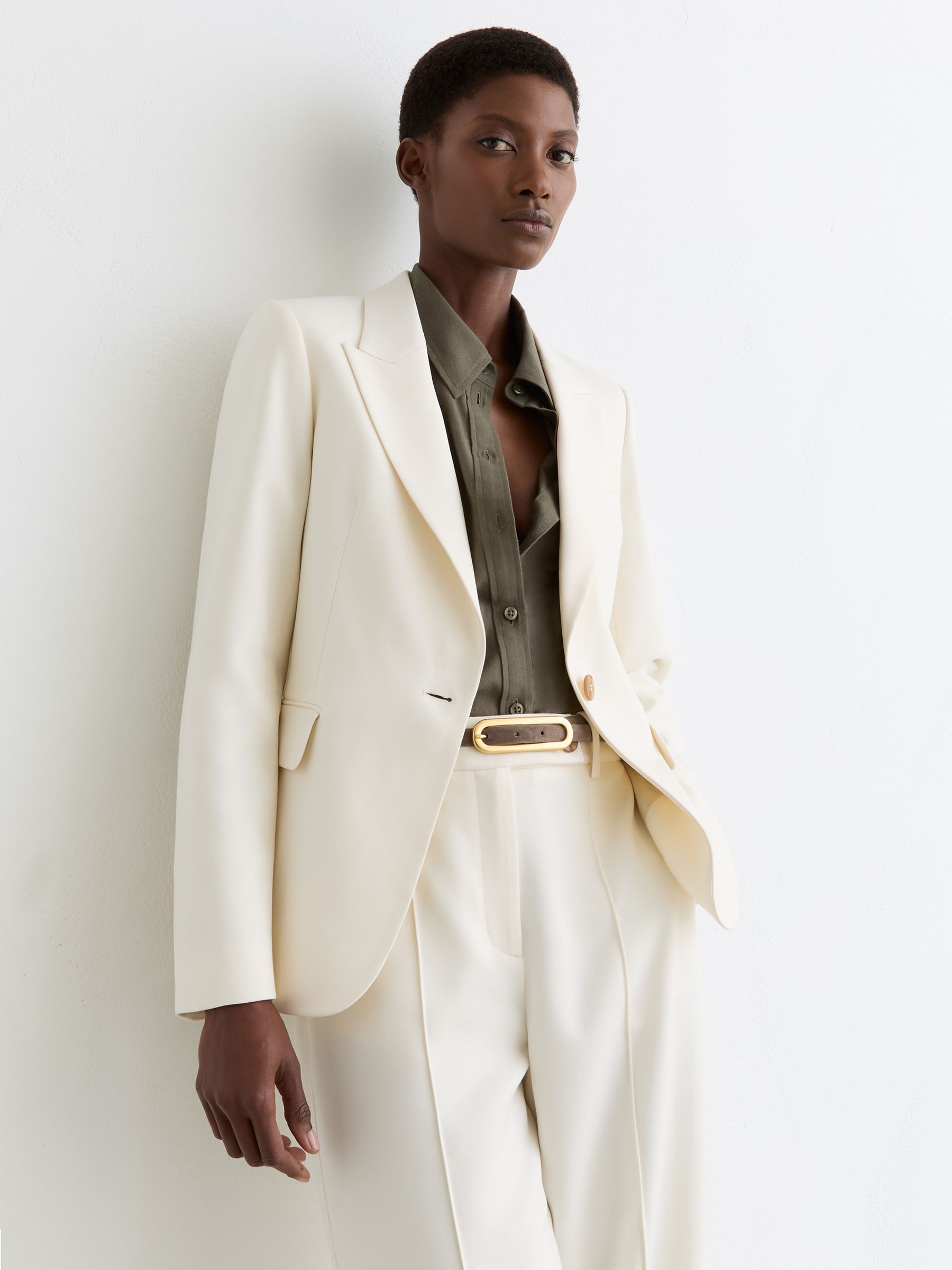 A woman wearing a cream tailored blazer with peak lapels, flap pockets, and a single button, paired with a dark green shirt.