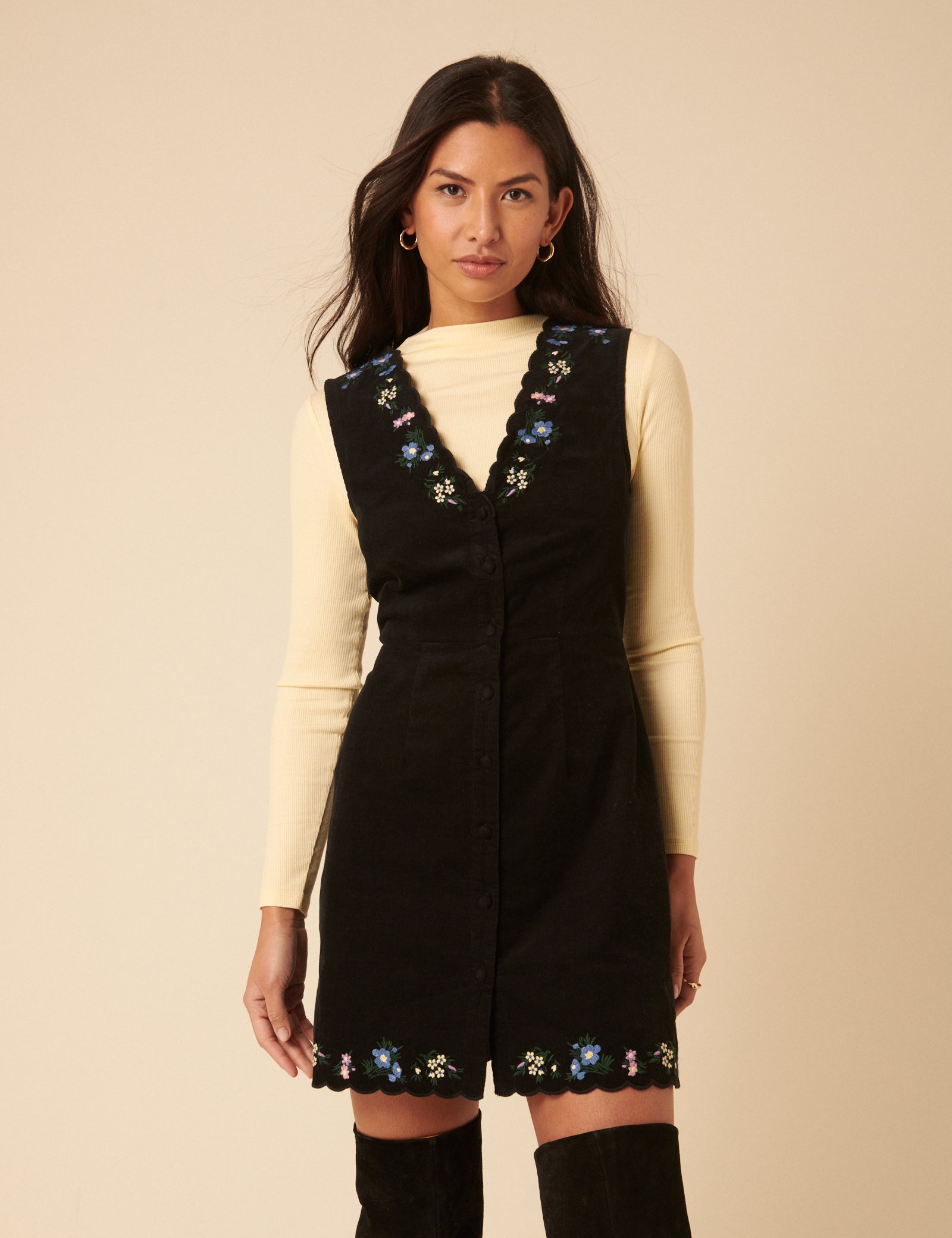 A woman wearing a black embroidered cord mini dress with floral details on the V-neckline and hem, sleeveless design, and button front.