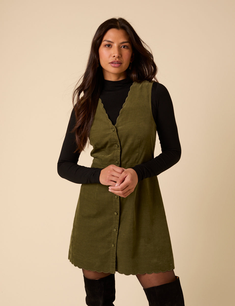 A woman wearing a green cord mini dress with scalloped V-neckline and button front, paired with a black long-sleeve top underneath.