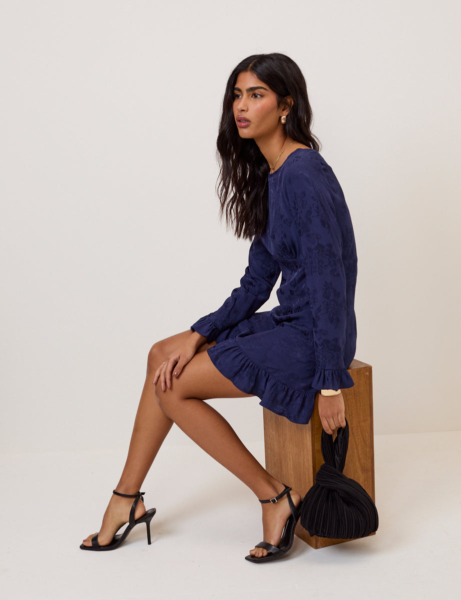 A woman wearing a blue floral satin jacquard mini dress with long sleeves, ruffled cuffs, and a ruffled hem, sitting on a wooden block with black high heels and holding a black pleated bag.