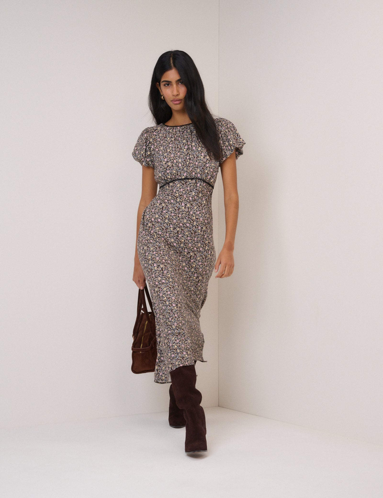 A woman wearing a black midi dress with a floral print, short puff sleeves, and a lace-trimmed skirt, standing against a plain background.