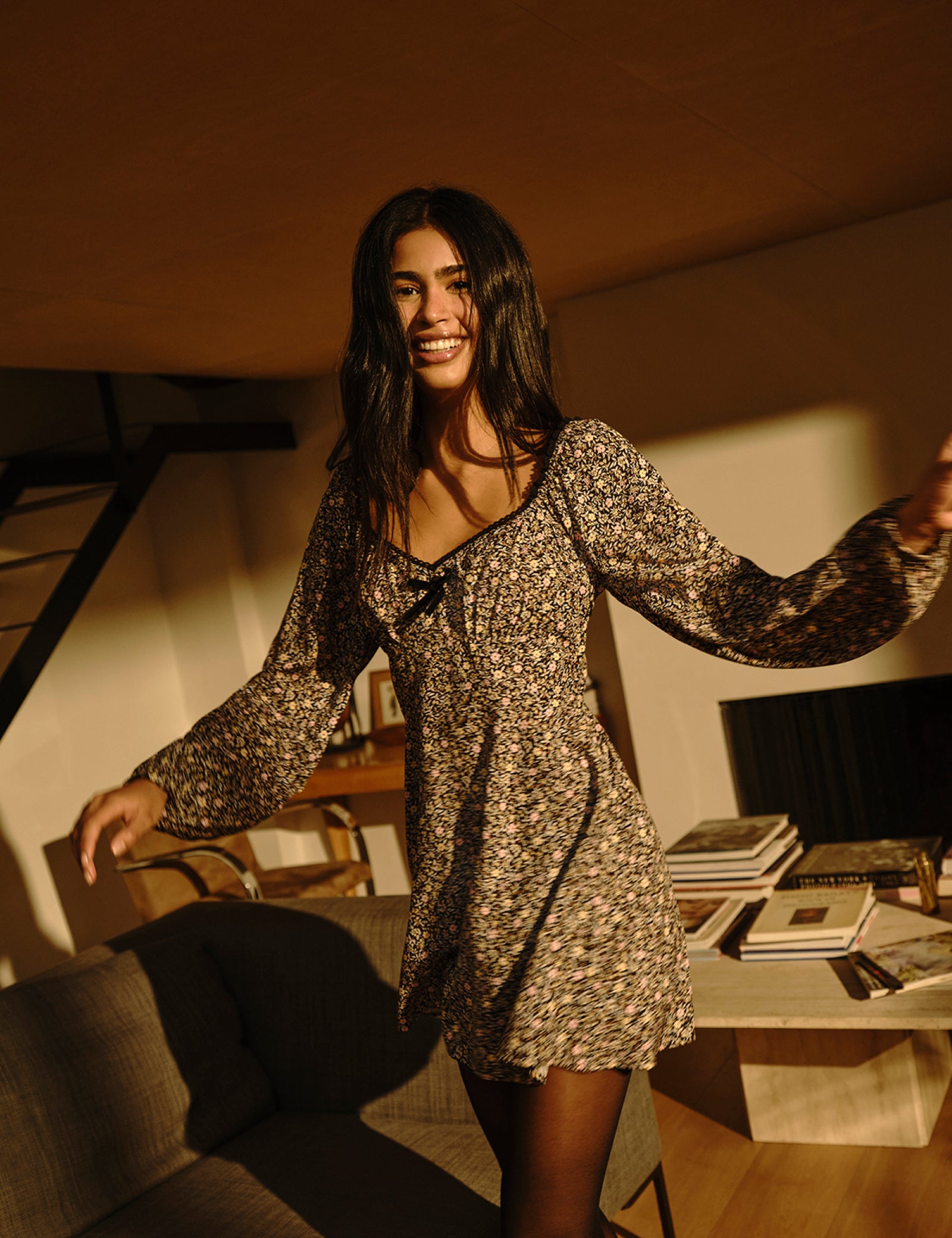 A woman wearing a floral mini dress with long balloon sleeves, a sweetheart neckline with lace trim, and a velvet bow, standing in a warmly lit room.