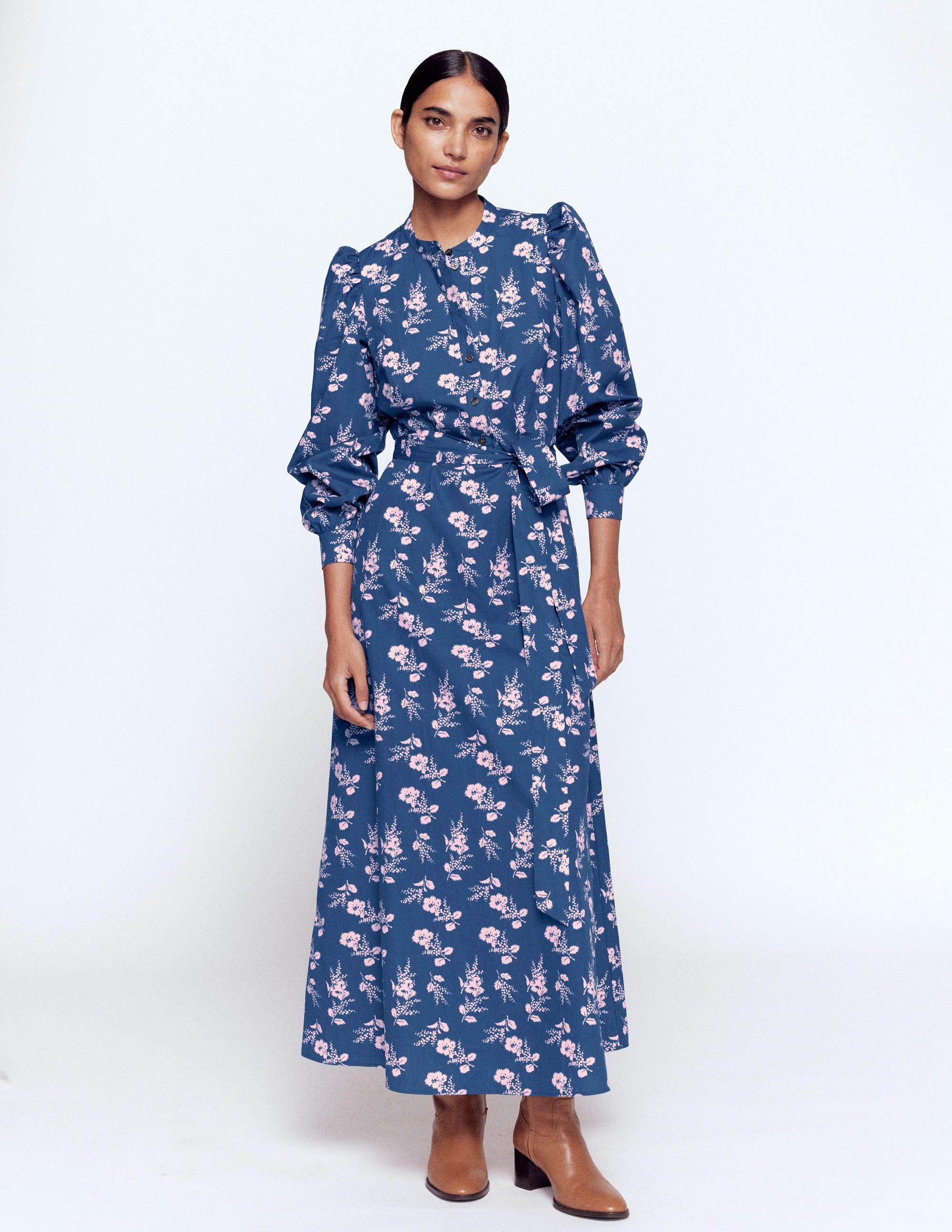 A woman wearing a navy floral midi dress with puffed shoulders, a waist tie, and side seam pockets, paired with tan ankle boots.