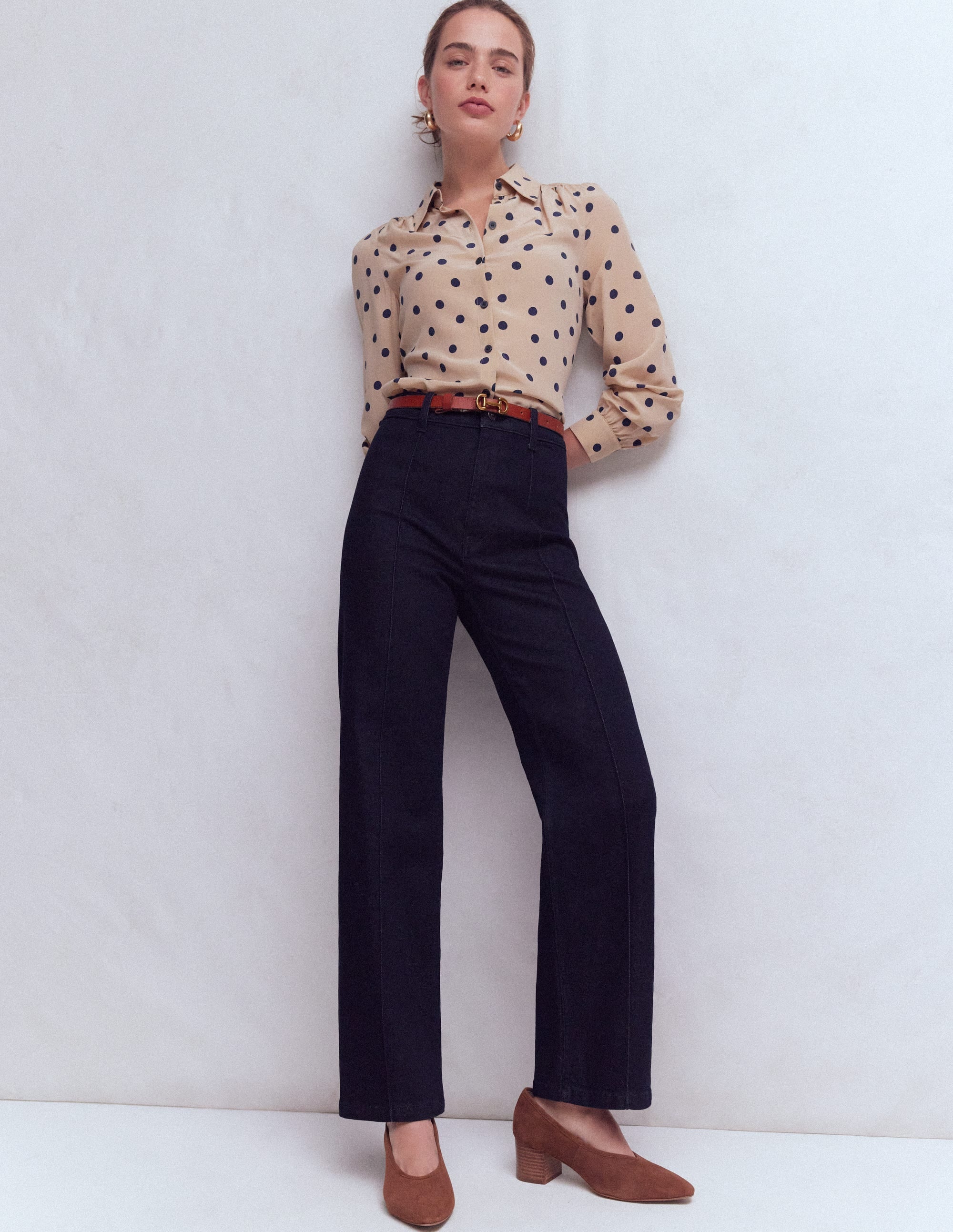 A woman wearing indigo rinse pintuck column leg jeans with a beige polka dot blouse, standing against a plain white background.