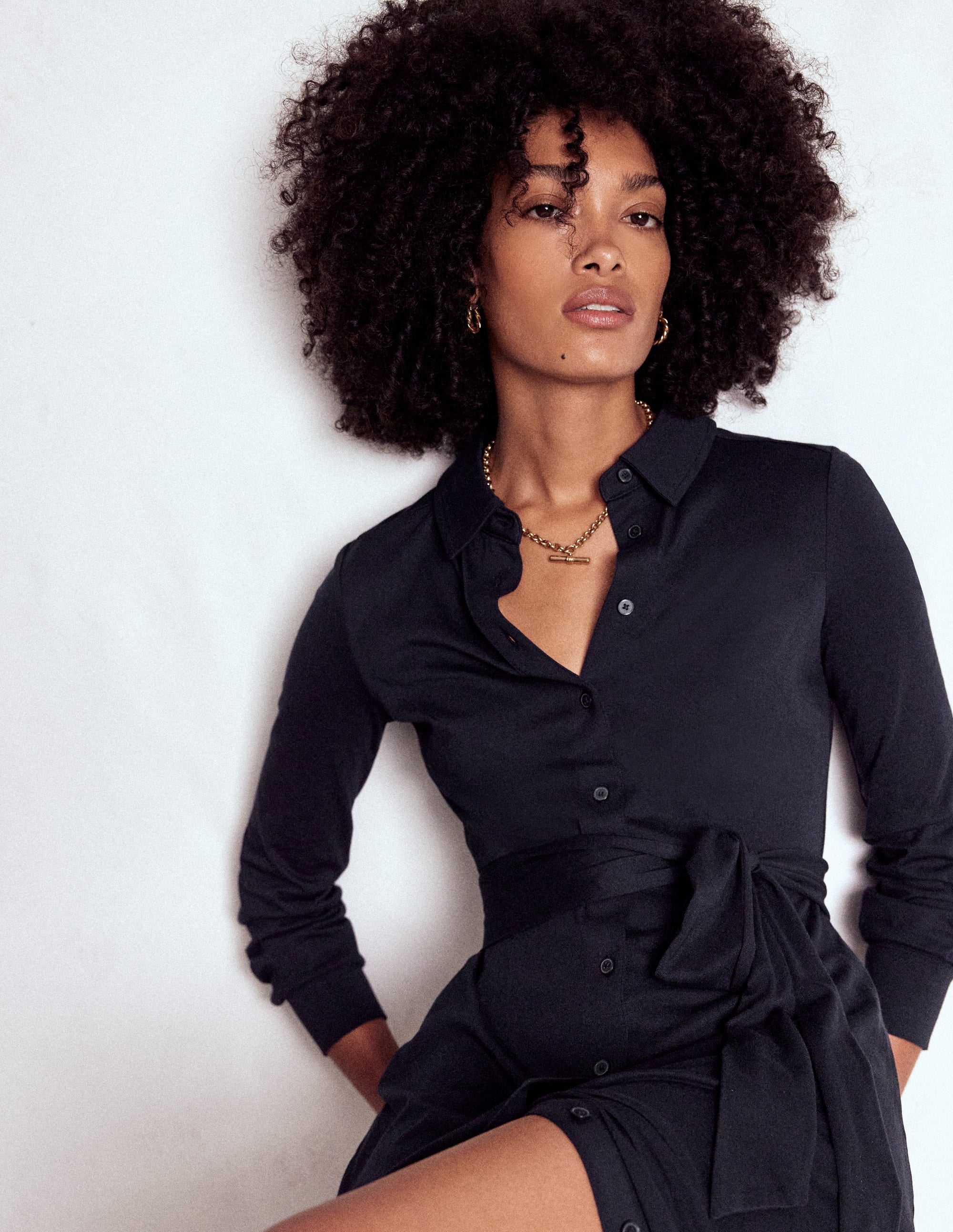 A woman wearing a navy shirt dress with a shirt collar, waist belt, and side pockets, standing against a white background.