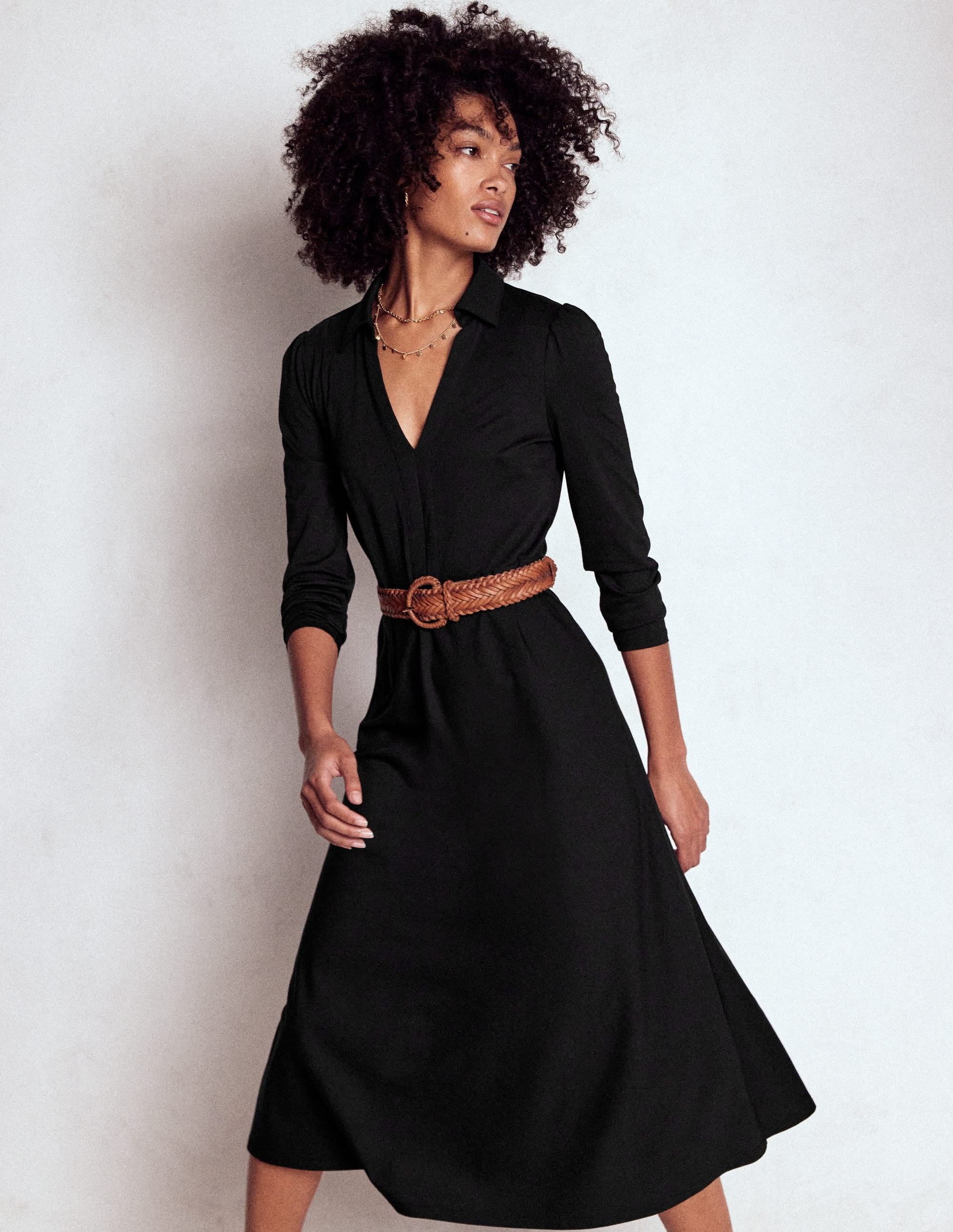 A woman wearing a black collared jersey dress with long sleeves and a flared skirt, accessorized with layered necklaces and a brown woven belt.