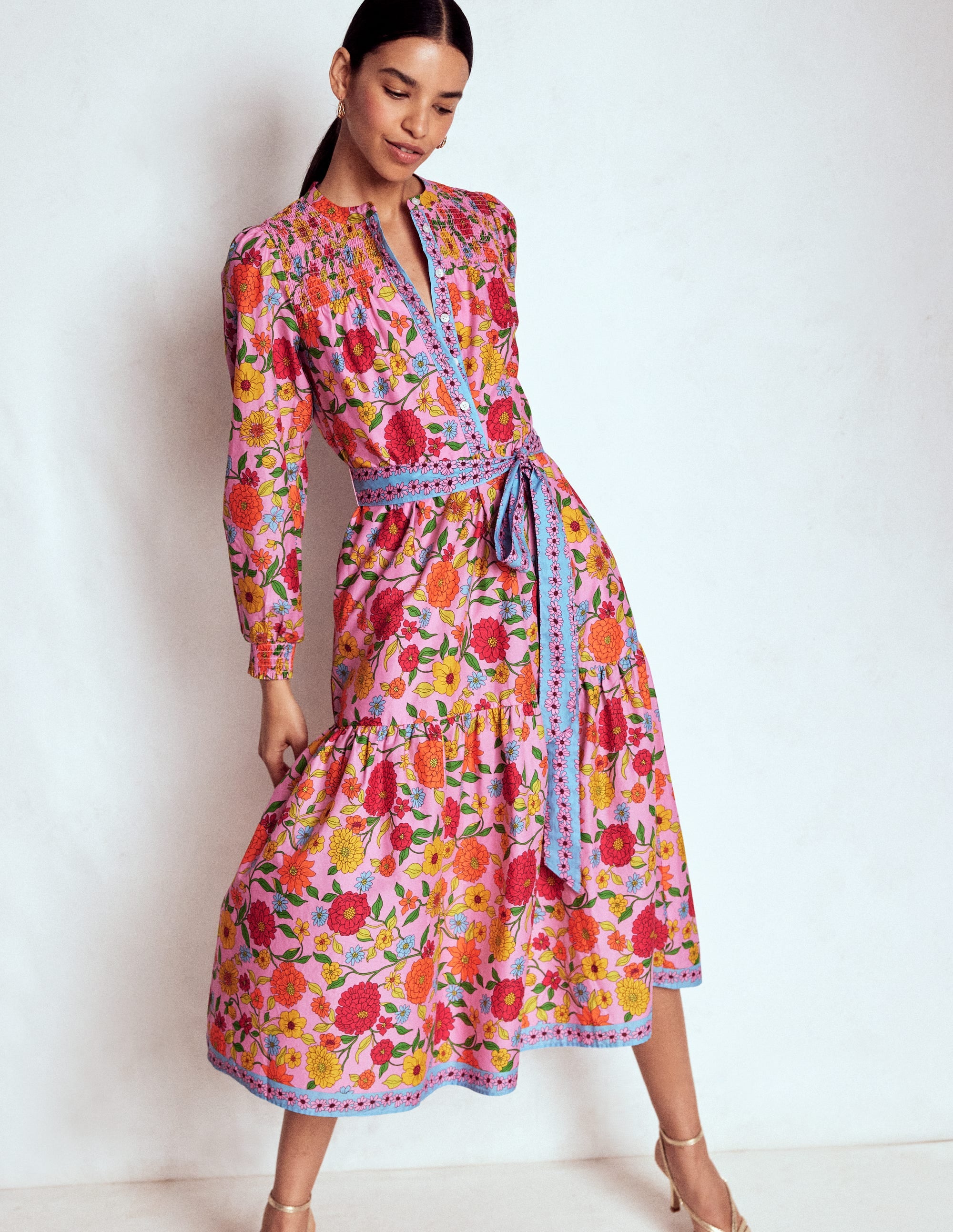 A woman wearing a colorful, floral-patterned midi dress with long sleeves, a fitted bodice, and a flared skirt, standing against a plain background.