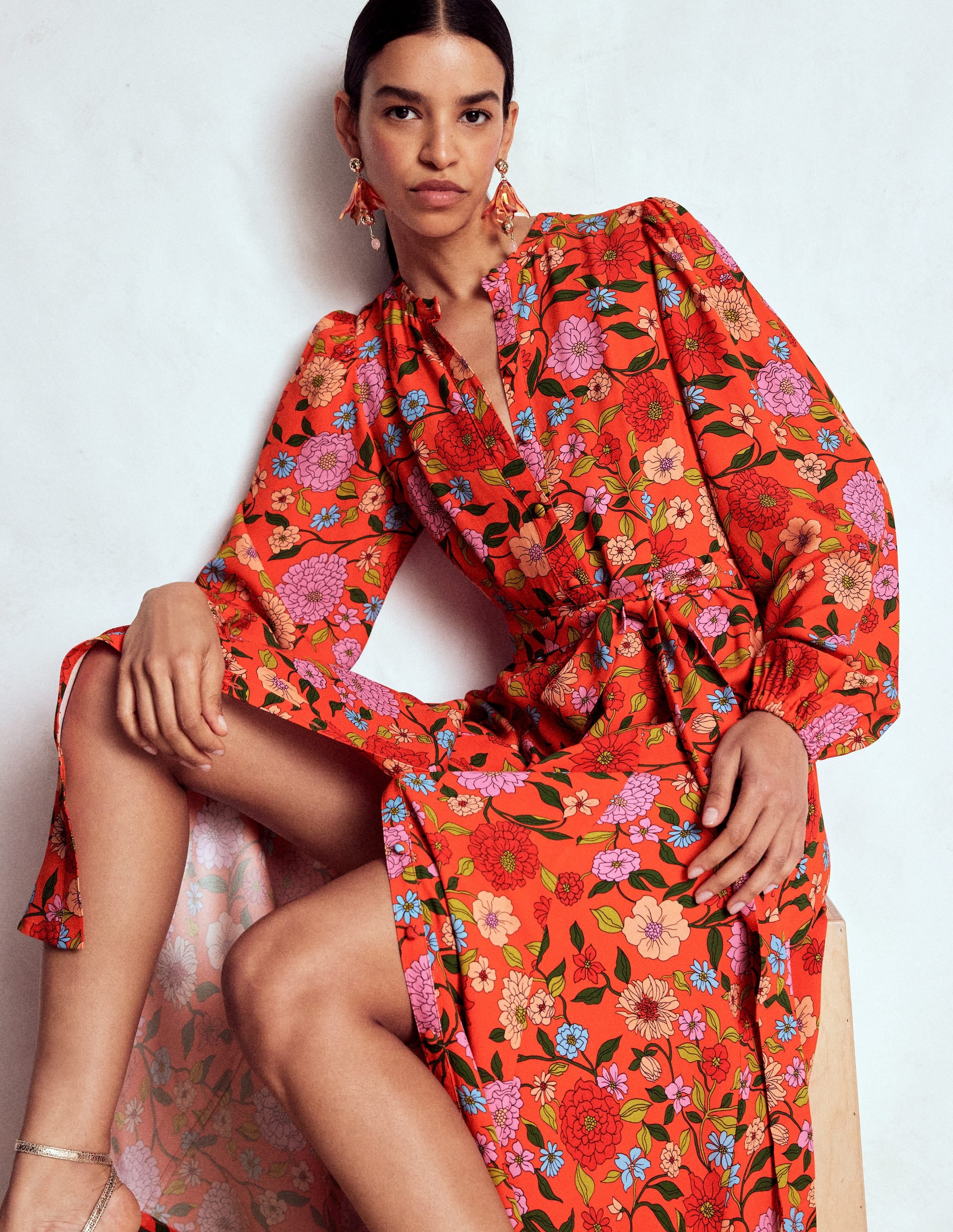 A woman wearing a long, floral maxi shirt dress in bright red with pink, purple, and blue flowers, featuring blouson sleeves, a tie belt, and pockets.