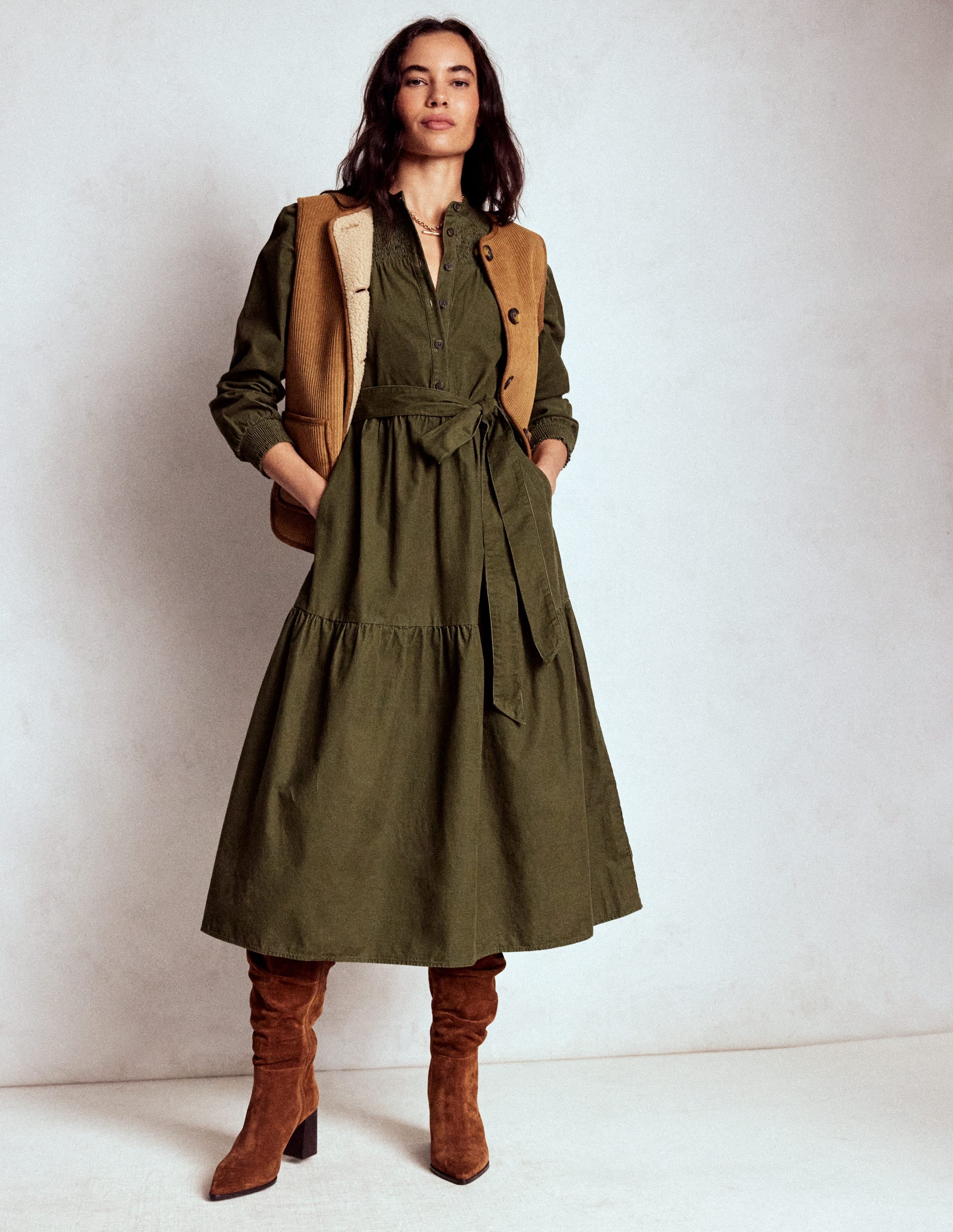 A woman wearing a woodland green midi dress with smocking, blouson sleeves, gathered tiers, and a self-fabric belt, paired with brown boots and a tan vest.