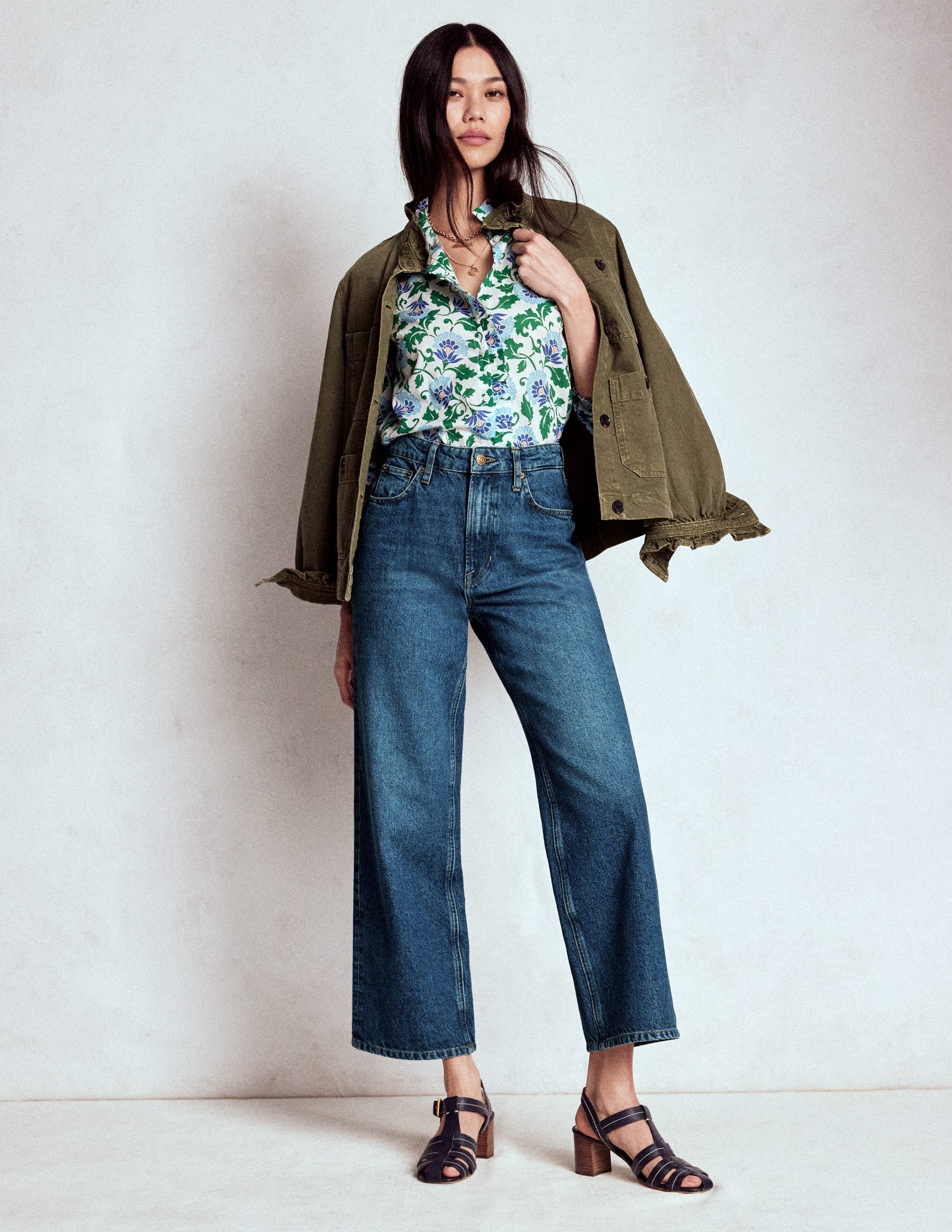 A woman wearing high-rise straight crop jeans in a medium blue wash with a high waist and straight legs, paired with a floral blouse and an olive green jacket.