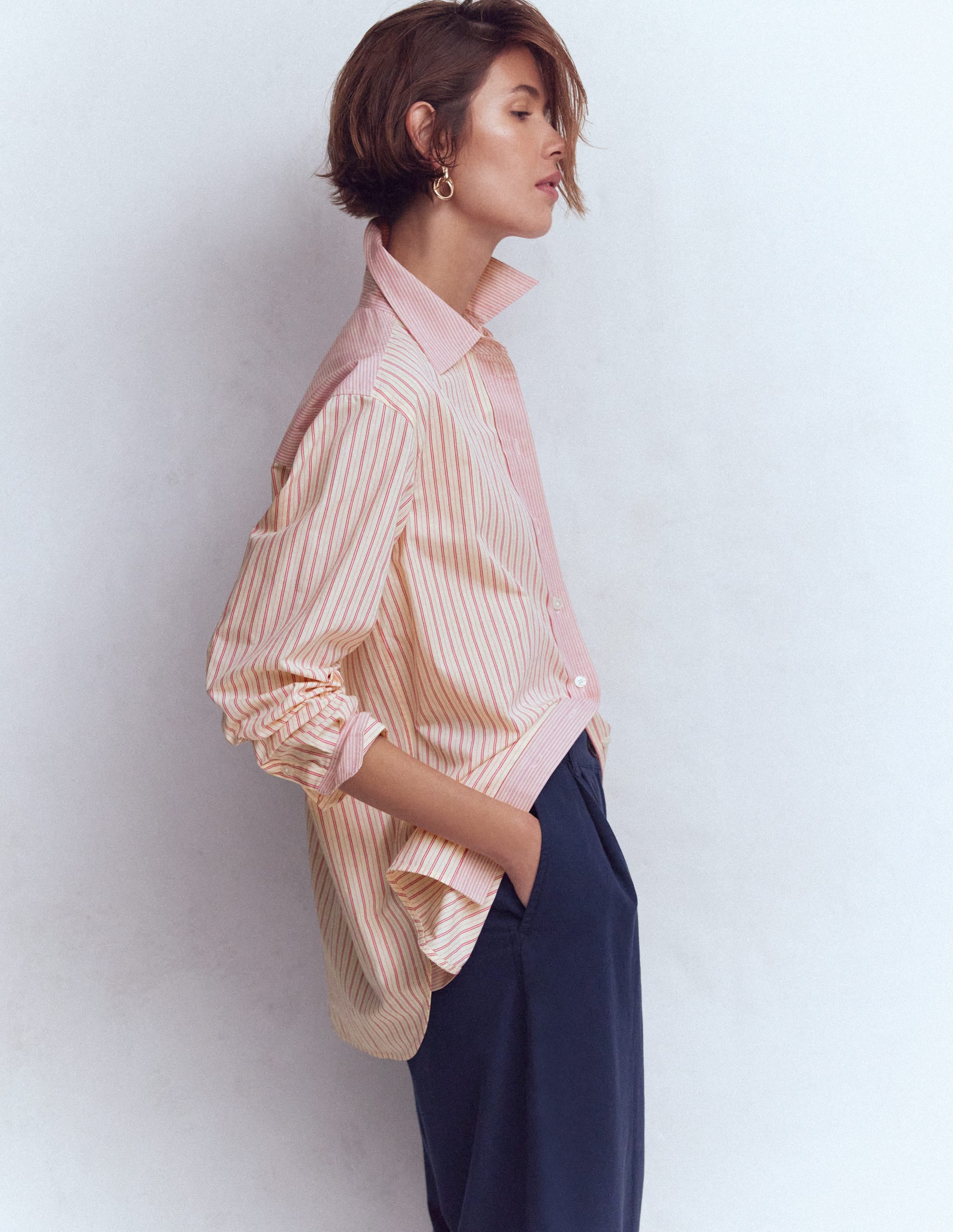 A woman wearing a pink and white striped cotton shirt with a relaxed fit, standing sideways with her hands in her pockets.