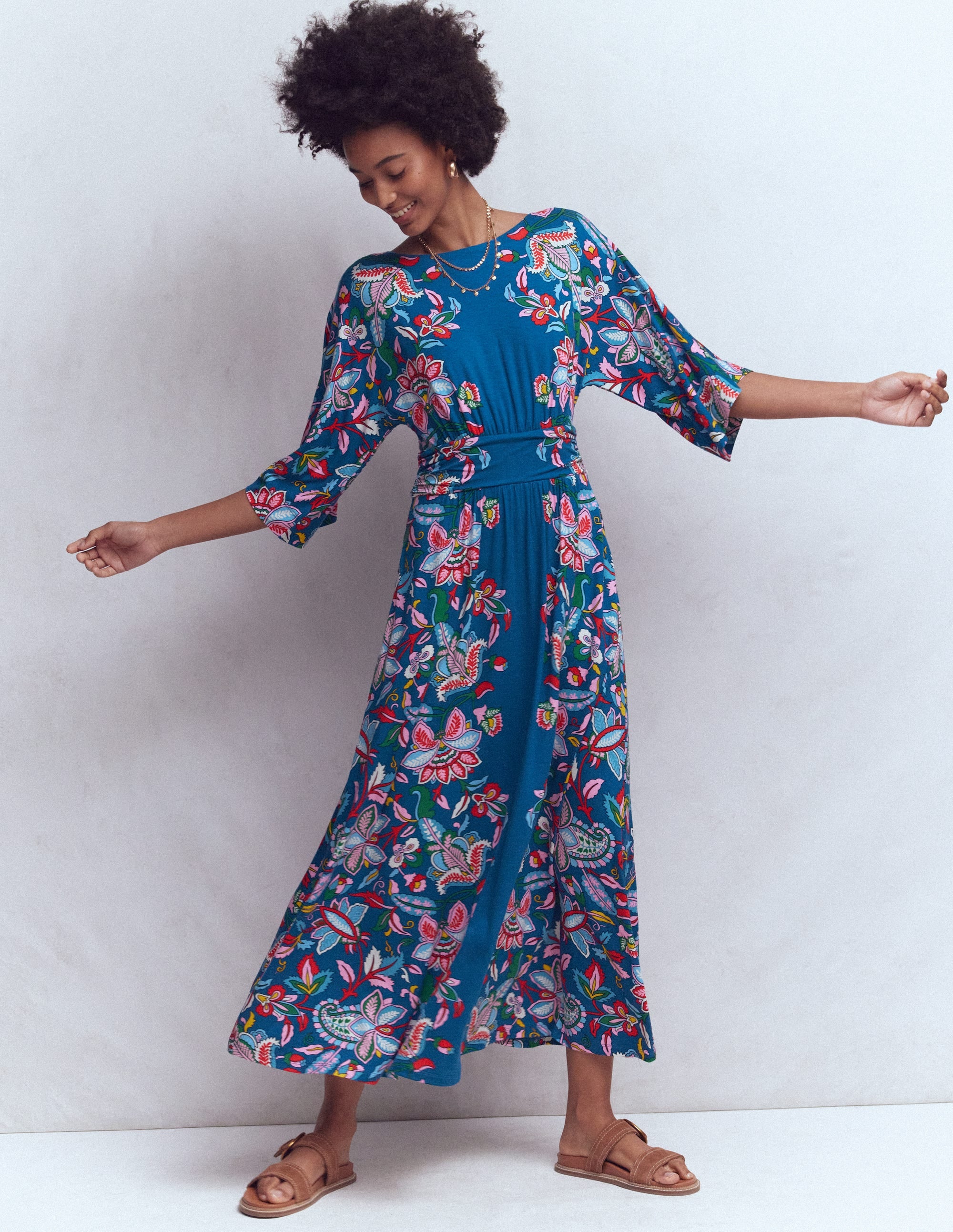 A woman wearing a blue midi dress with a vibrant botanical print, wide sleeves, and a ruched waistband, standing against a plain background.