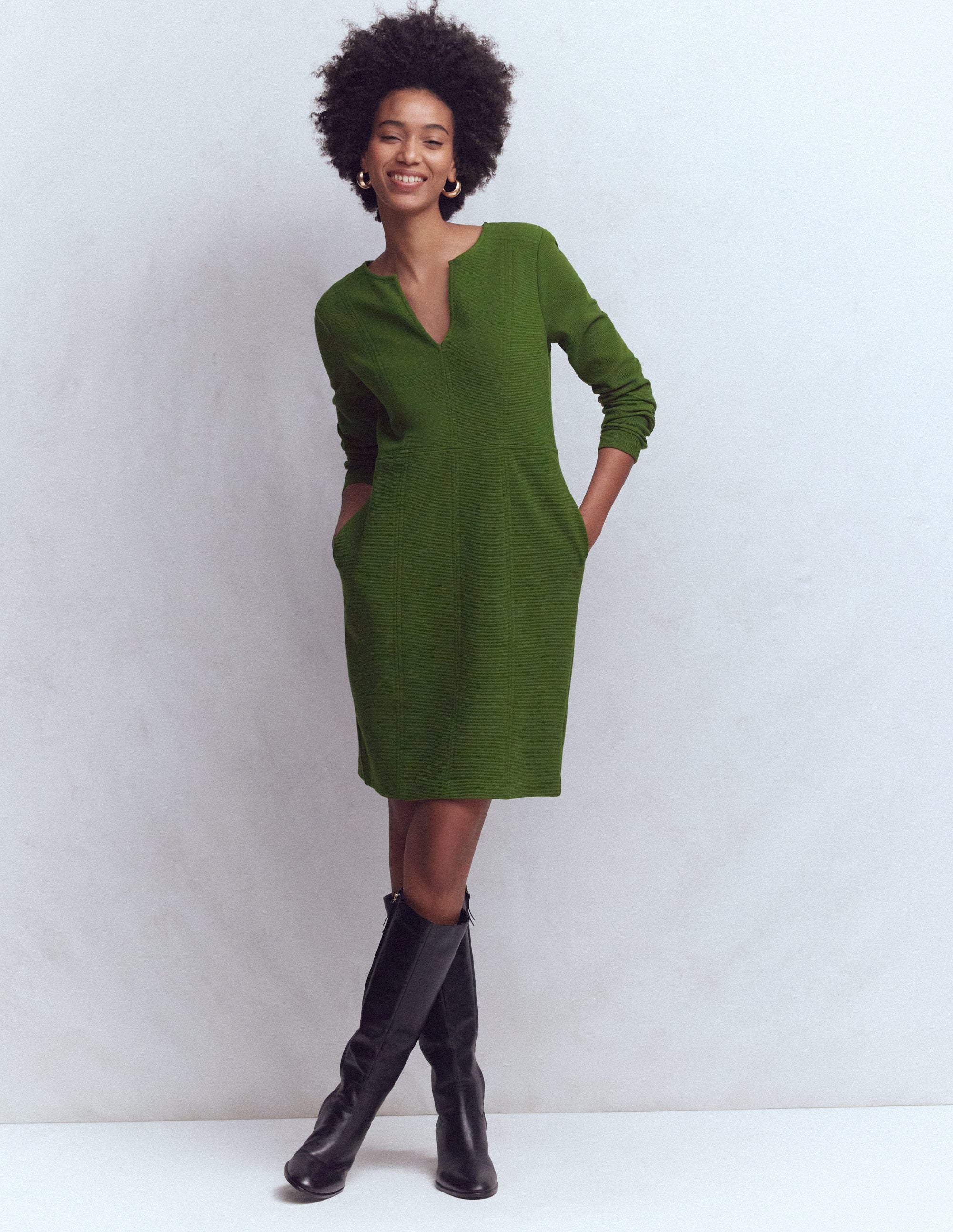 A woman wearing a green shift dress with a notch neckline, long sleeves, and side pockets, standing against a plain background.