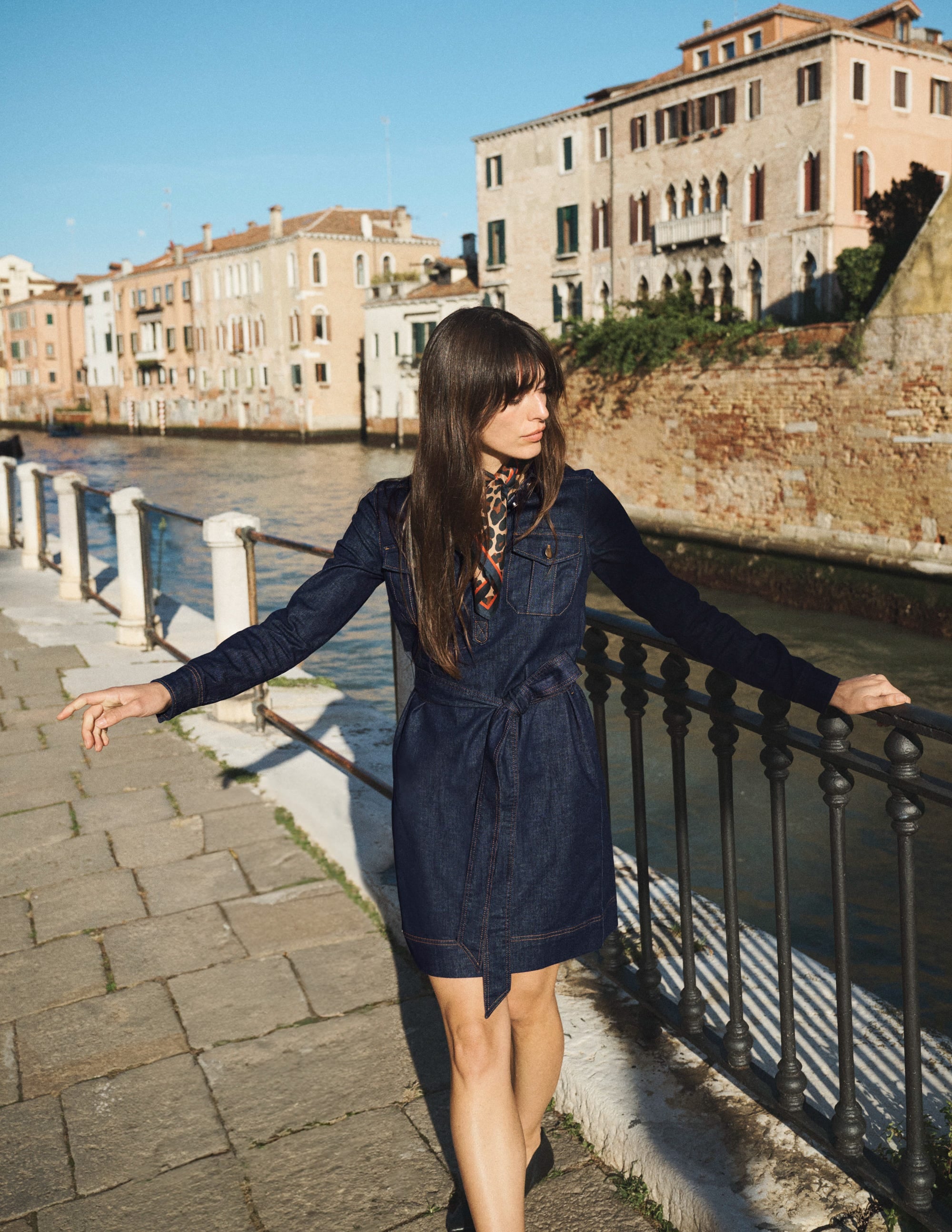 A woman wearing a dark indigo denim dress with a half placket, gold buttons, and a self-tie belt, standing by a canal with historic buildings in the background.