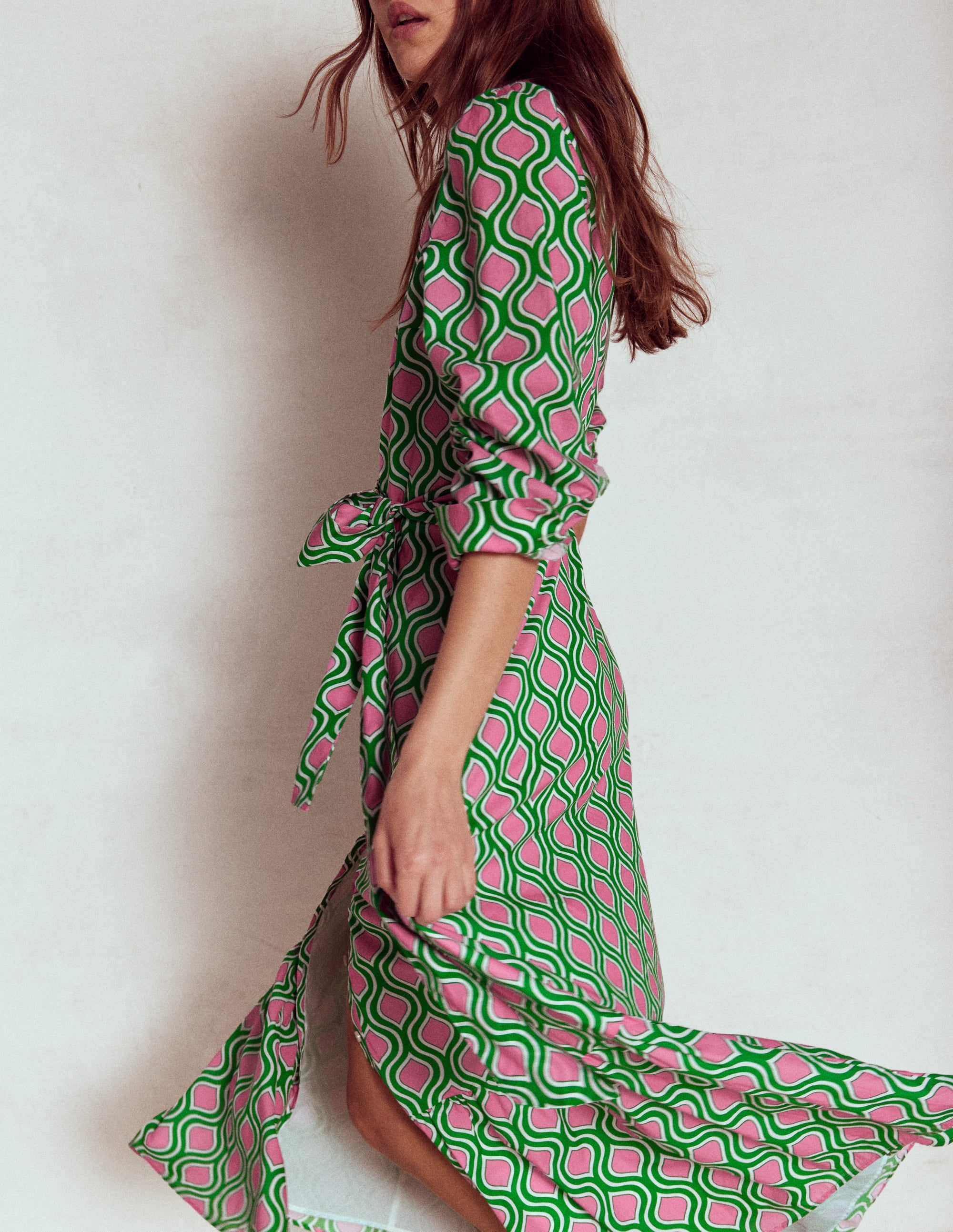 A woman wearing a green and pink geometric print tiered shirt dress with a fabric tie belt and button cuffed sleeves.