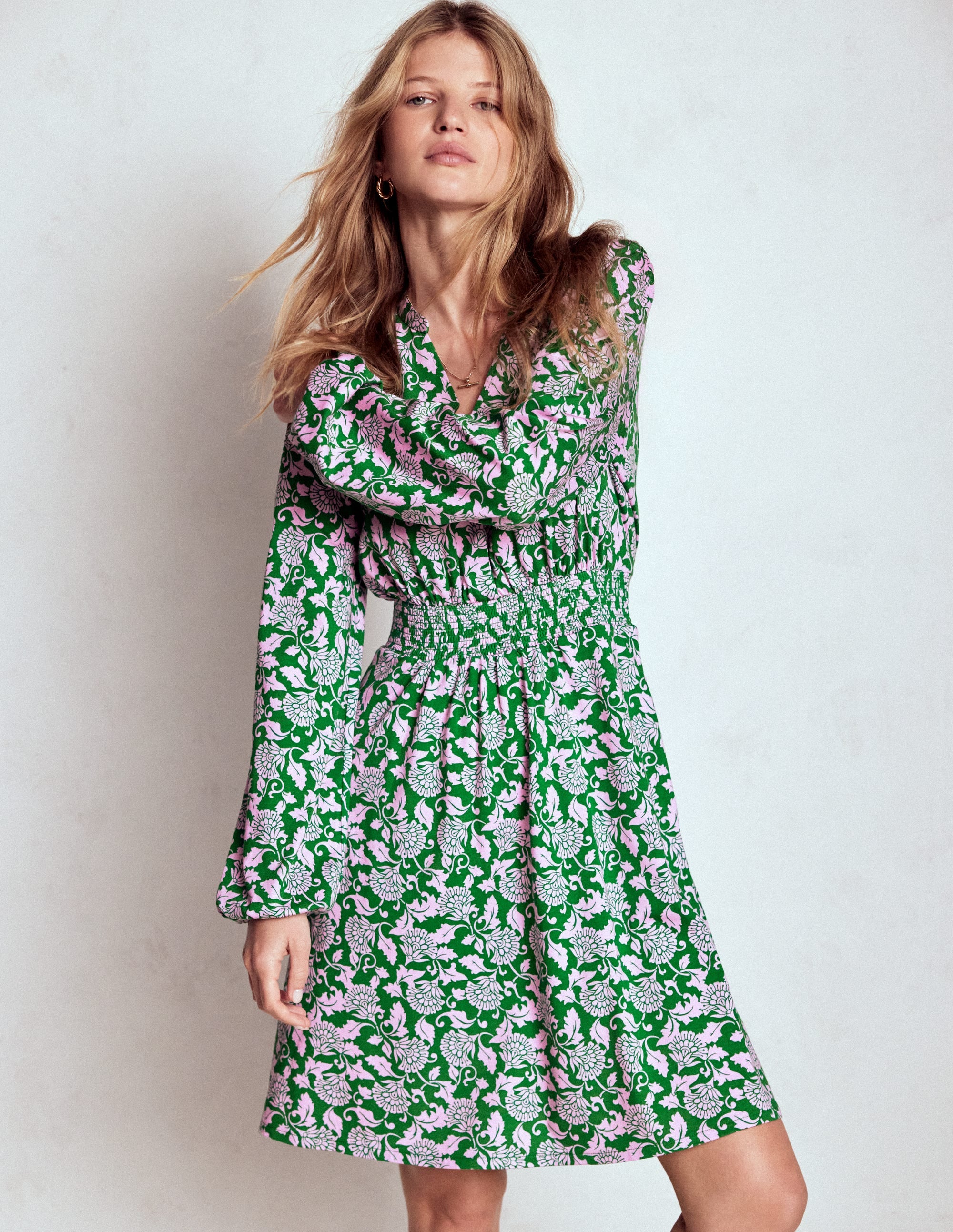 A woman wearing a knee-length dress with a green, pink, and white ornate vine floral pattern, long sleeves, and a fit and flare silhouette.