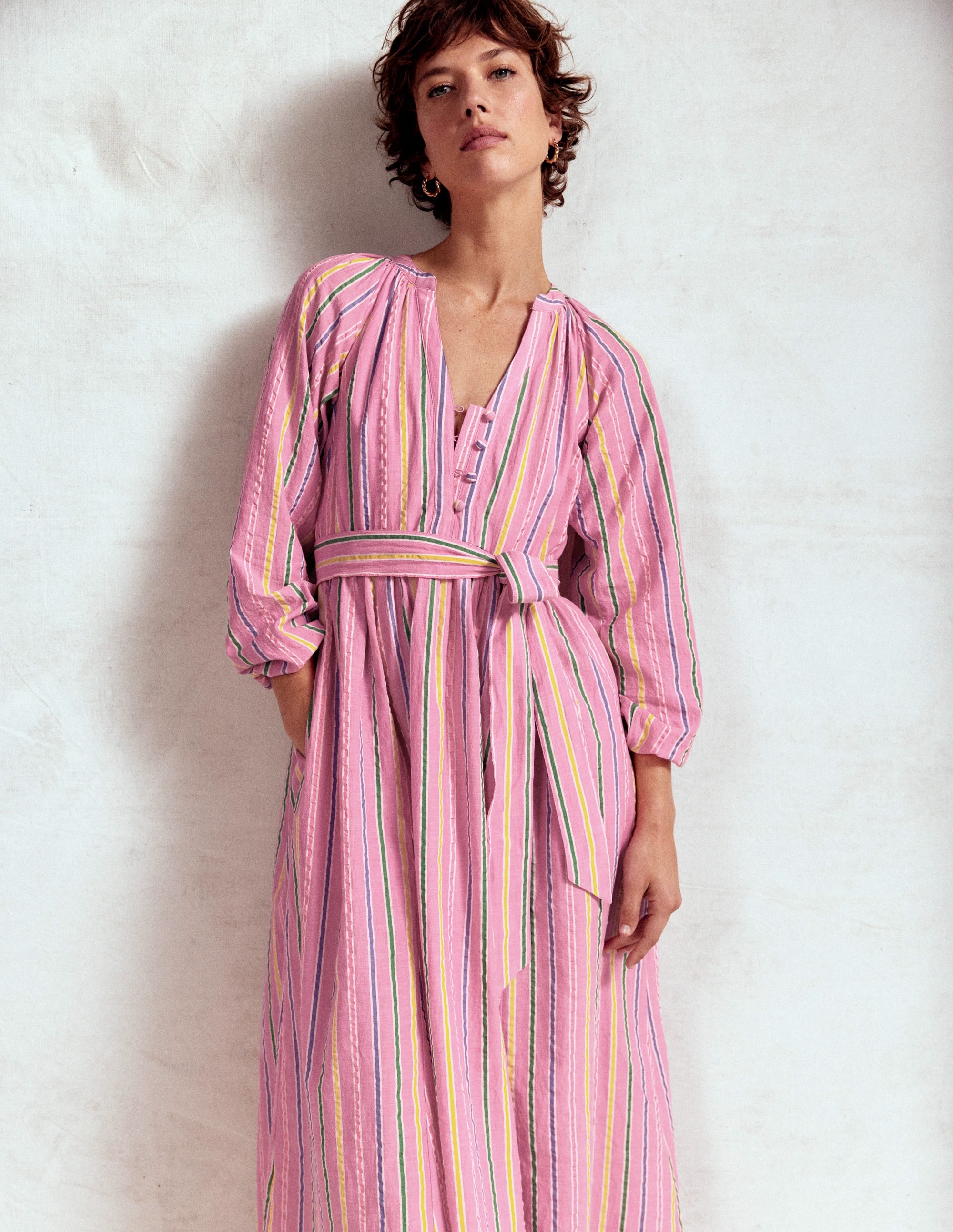 A woman wearing a sorbet pink maxi dress with seersucker stripe pattern, puff sleeves, and a self-tie belt.