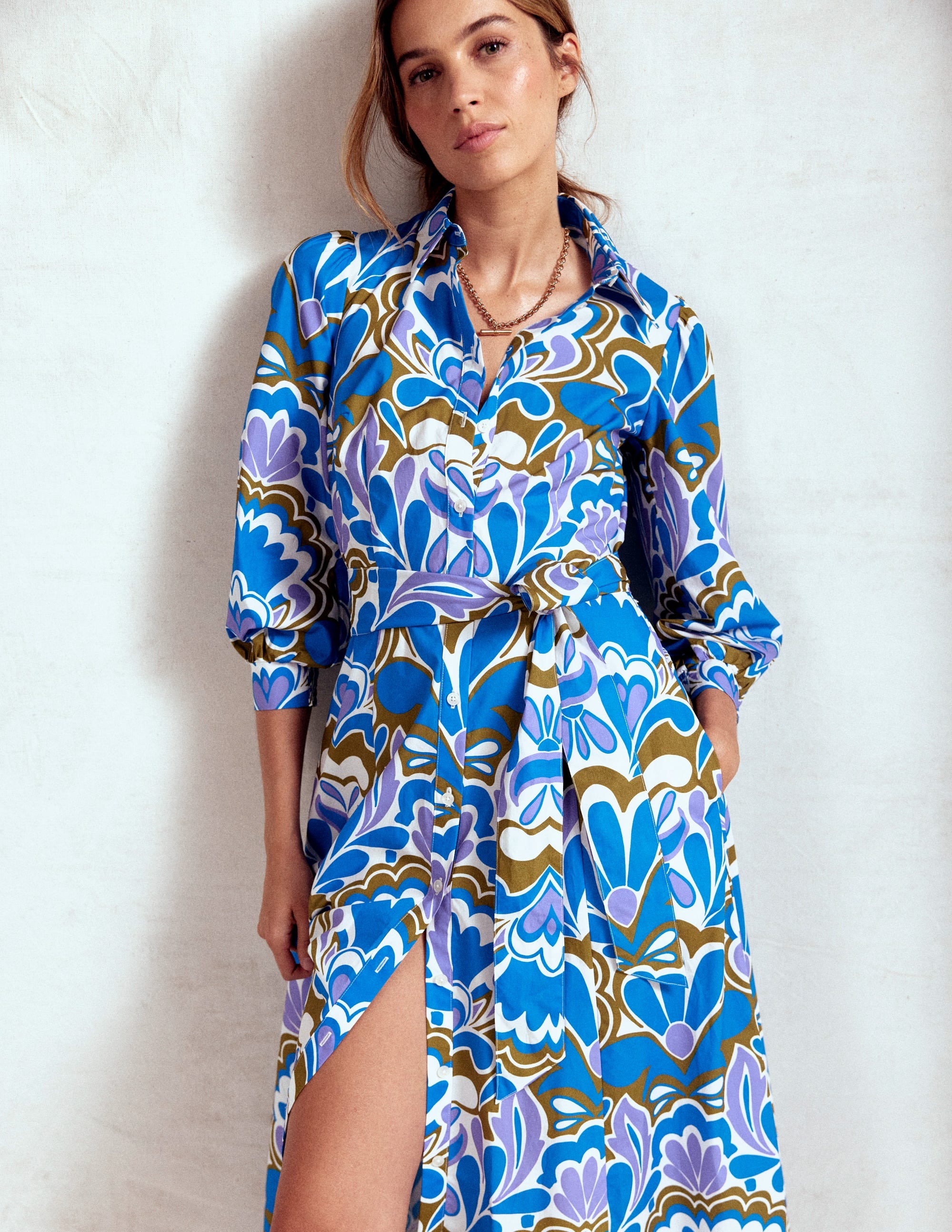 A woman wearing a long, blue floral shirt dress with a waist belt and side pockets, standing against a plain background.